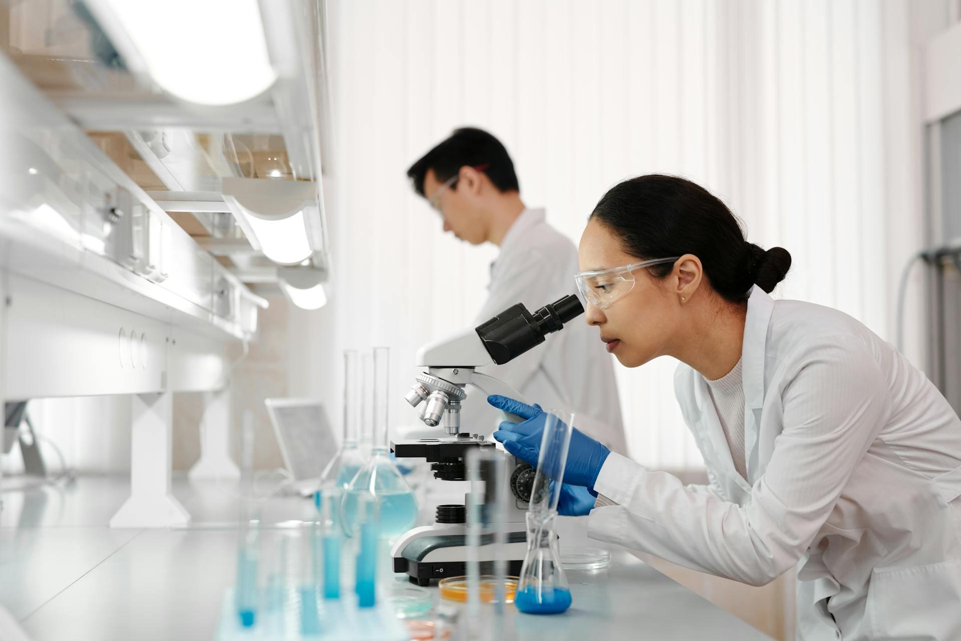 Persoon met beschermbril in witte jas en met blauwe handschoenen kijkt in microscoop