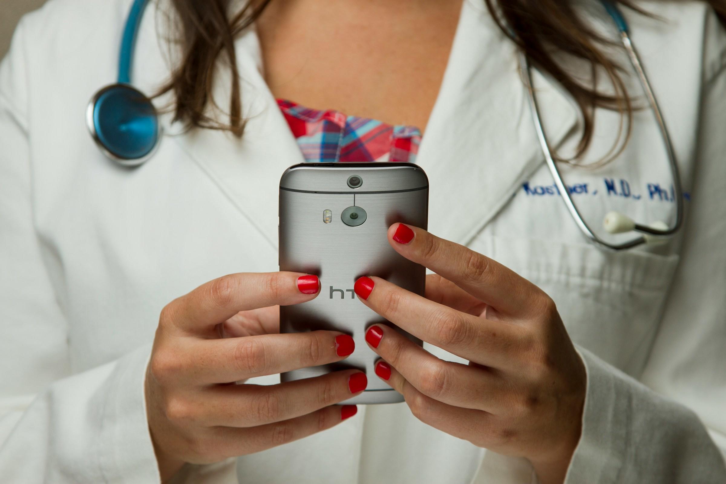 Persoon met witte jas en stethoscoop om houdt met beide handen IPhone vast