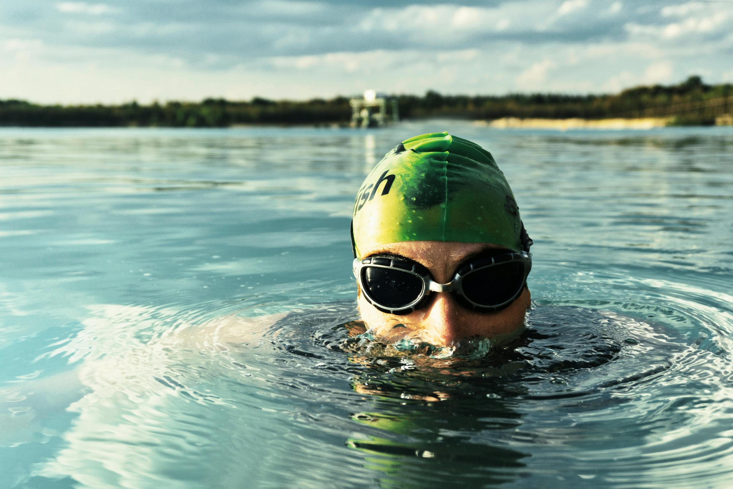 Een zwemmer met een groene badmuts en zwarte zwembril komt boven het wateroppervlak in een buitenwater.