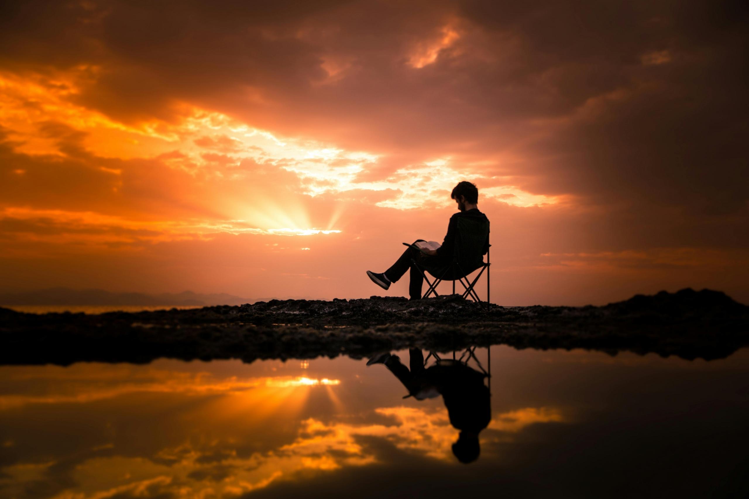 Een persoon zit op een klapstoel en leest een boek tijdens een zonsondergang, met reflecties in het water.