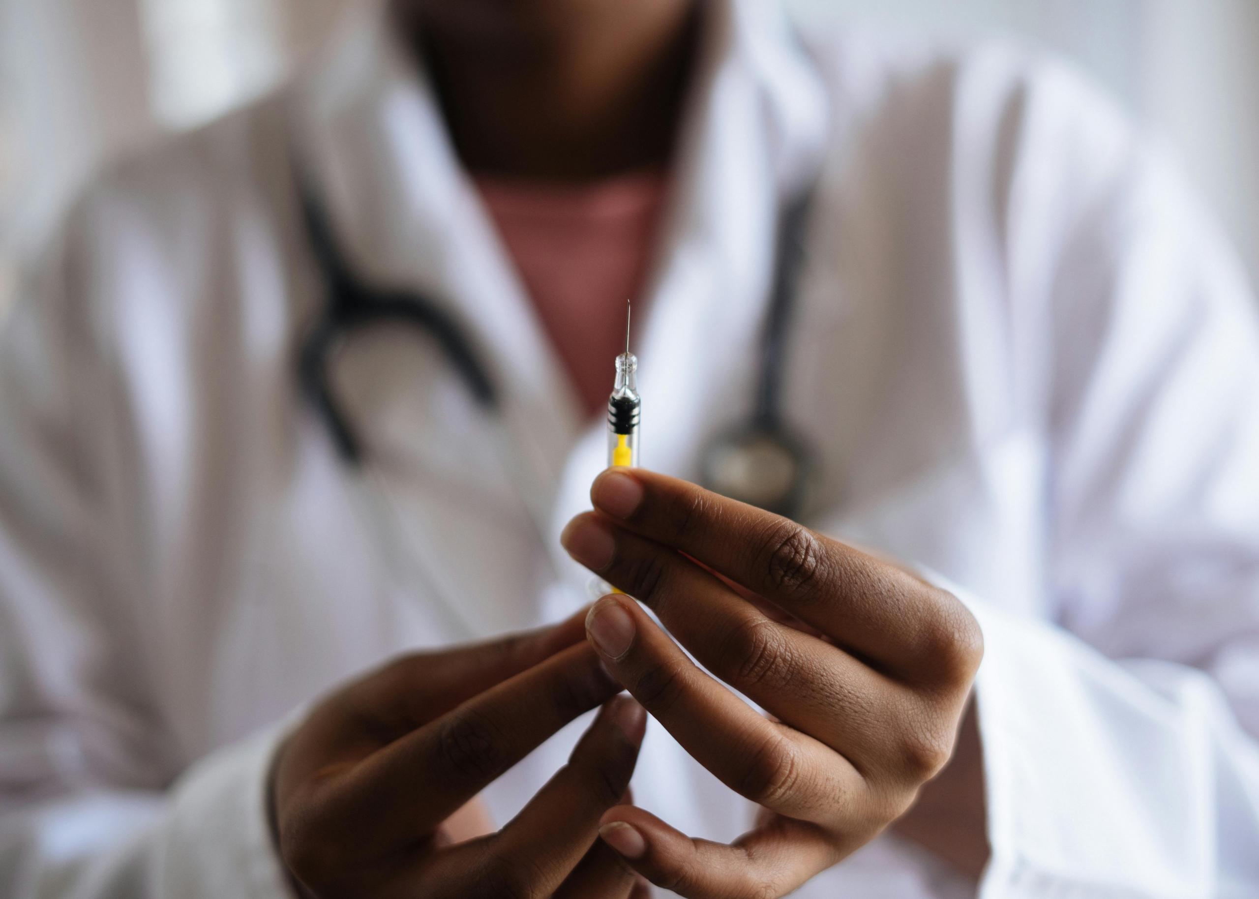 Een zorgprofessional in een witte jas die een injectiespuit vasthoudt. Op de achtergrond is een stethoscoop zichtbaar om de nek van de persoon.