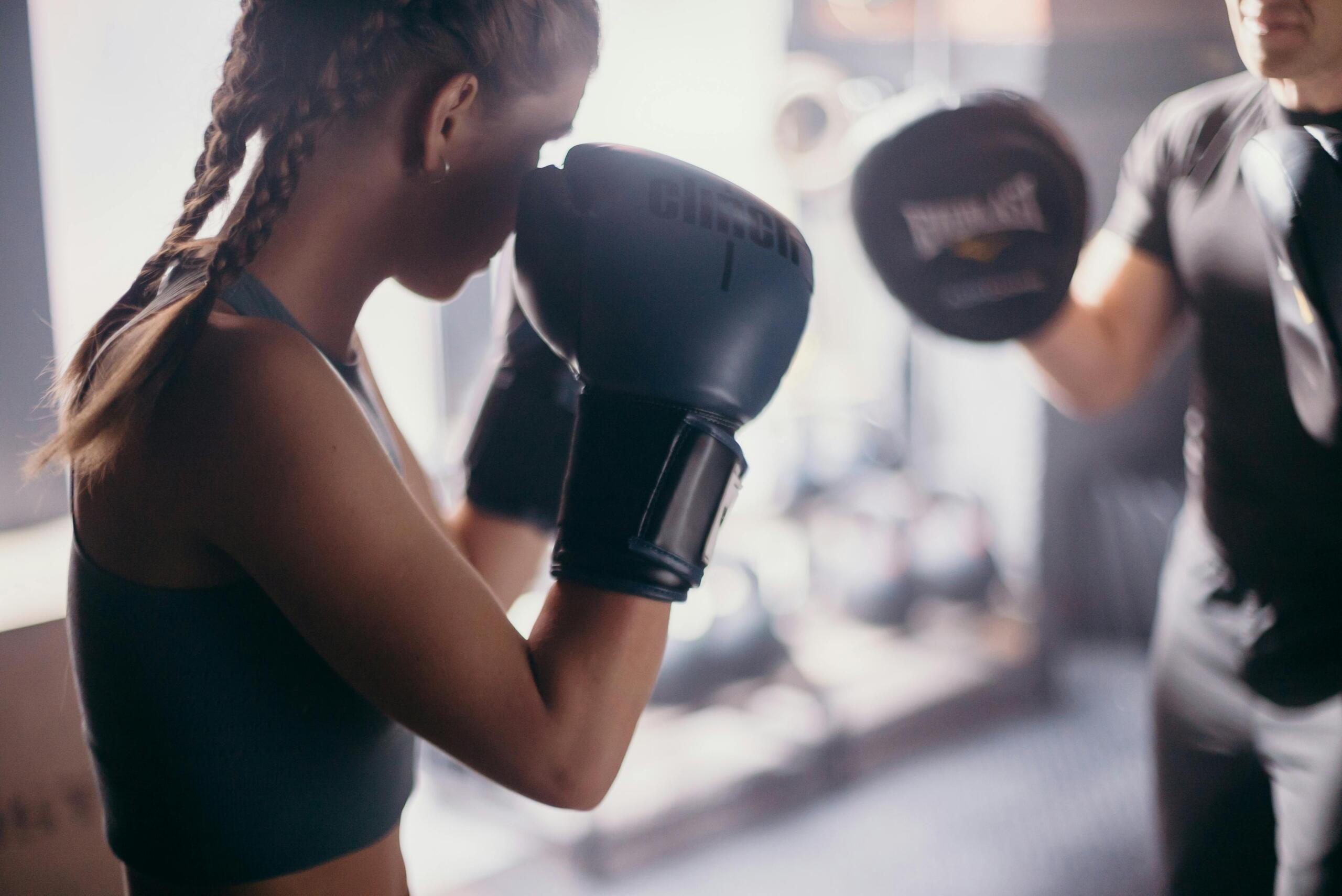 Een vrouw met bokshandschoenen traint met een coach die pads vasthoudt, terwijl ze zich concentreert op haar verdediging.