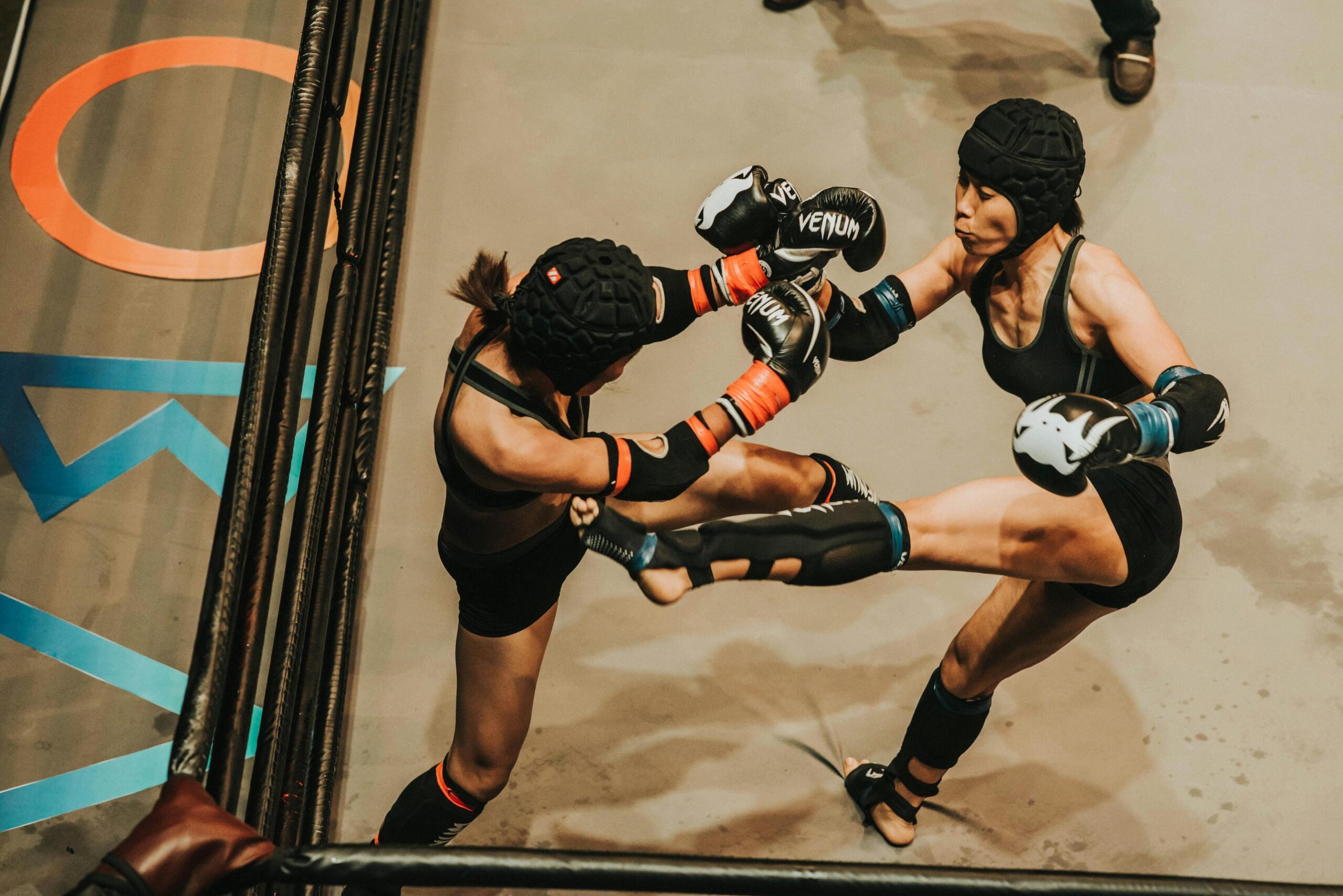 Twee vrouwelijke kickboksers in de ring, beide gelijktijdig een hoge trap uitvoerend, met beschermende uitrusting en intense focus.