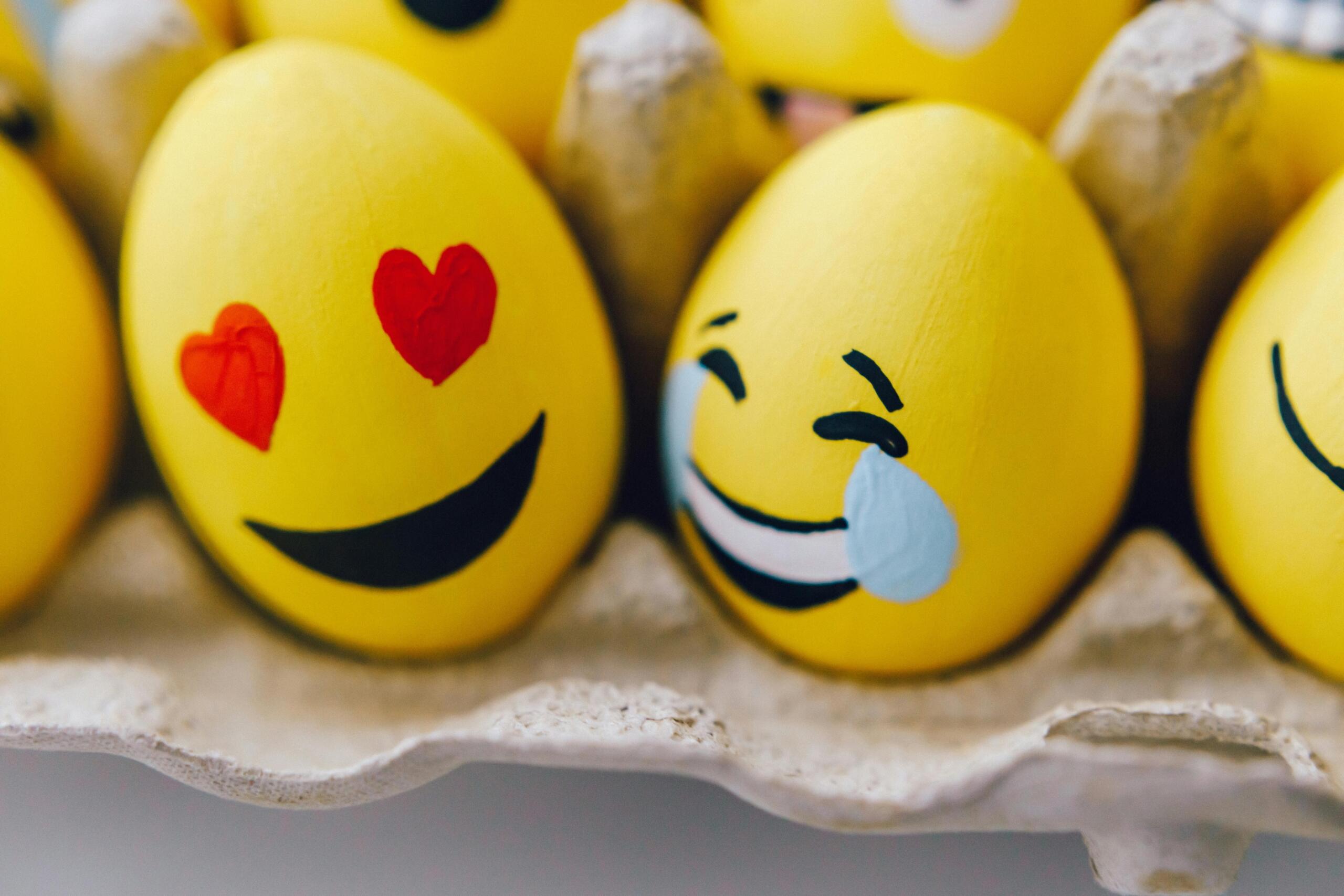 Gele eieren met geschilderde emoji-gezichtjes, waaronder een met hartvormige ogen en een met tranen van het lachen.