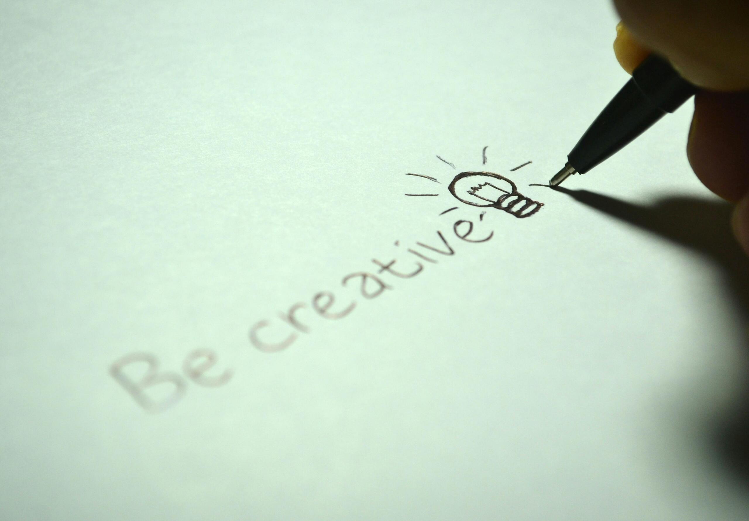 Een hand schrijft de woorden "Be creative" met een kleine tekening van een gloeilamp.