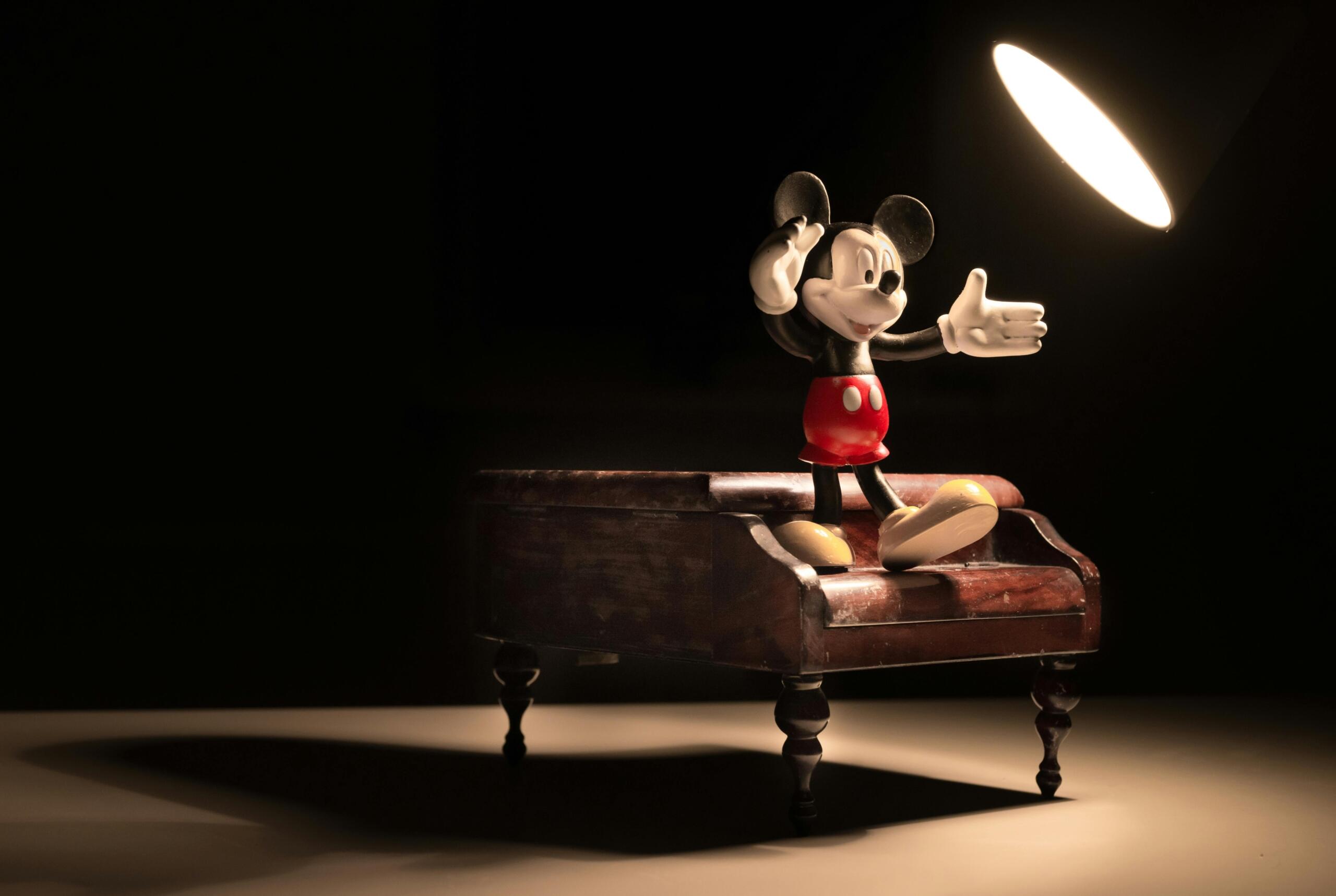 Een figuurtje van Mickey Mouse staat op een houten piano onder een felle spotlamp in een donkere ruimte.