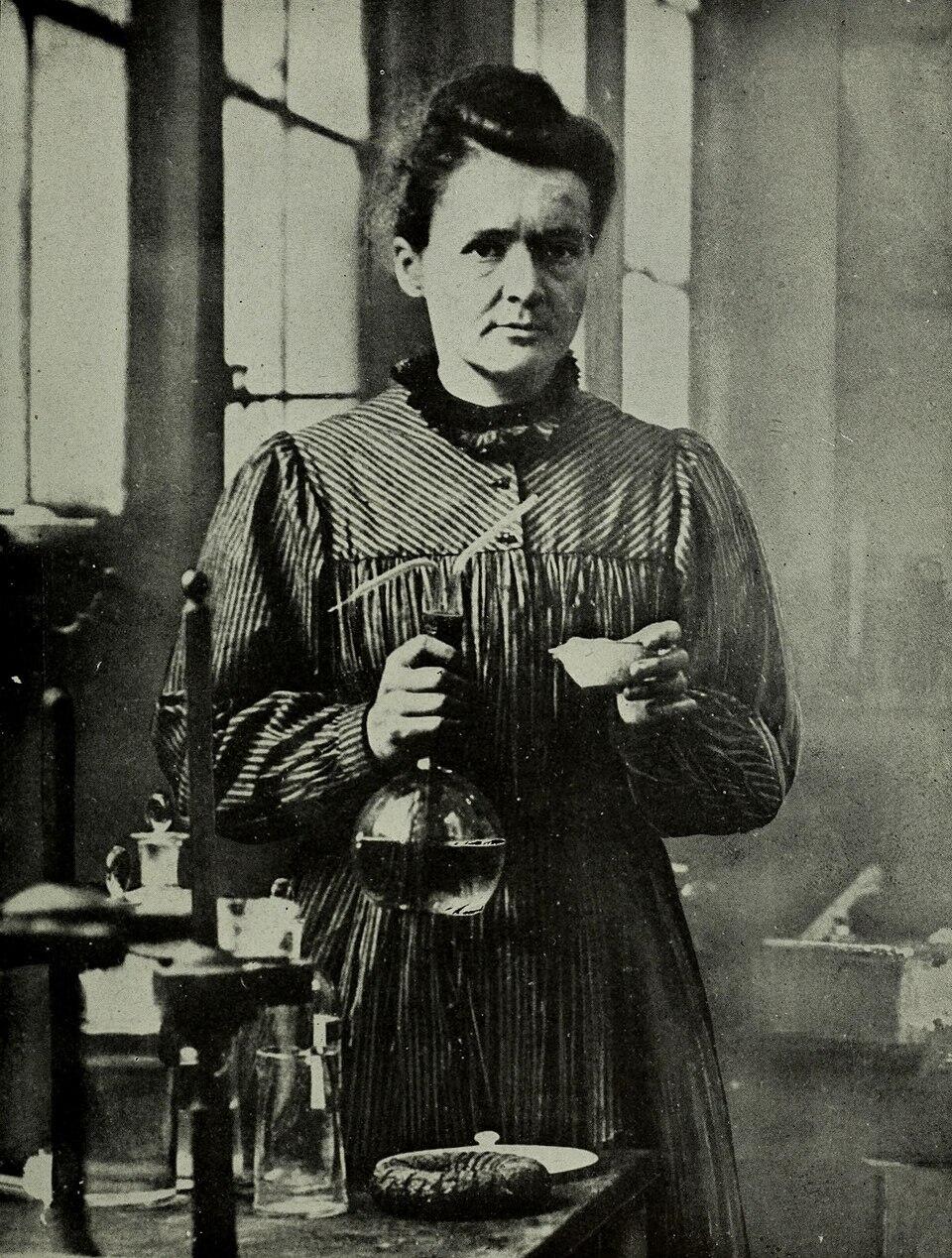 Portret van Marie Curie in een laboratorium, houdend een laboratoriumfles en kijkend naar de camera.