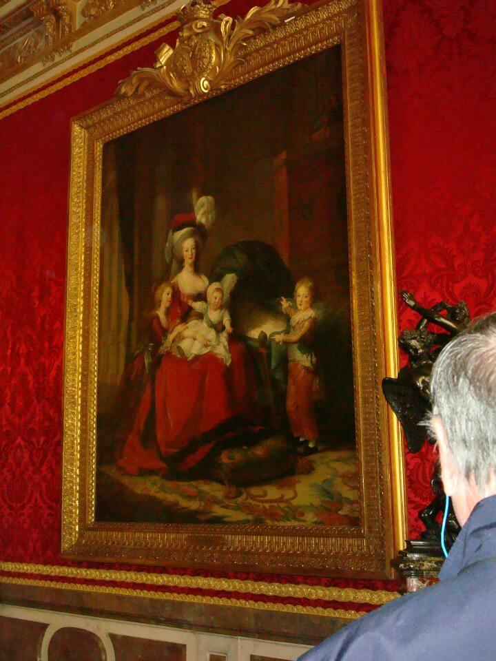 Marie Antoinette zit op een stoel, omringd door haar drie kinderen, in een intieme en moederlijke pose, gekleed in een rode jurk met een witte hoed, tegen een achtergrond van klassieke architectuur.