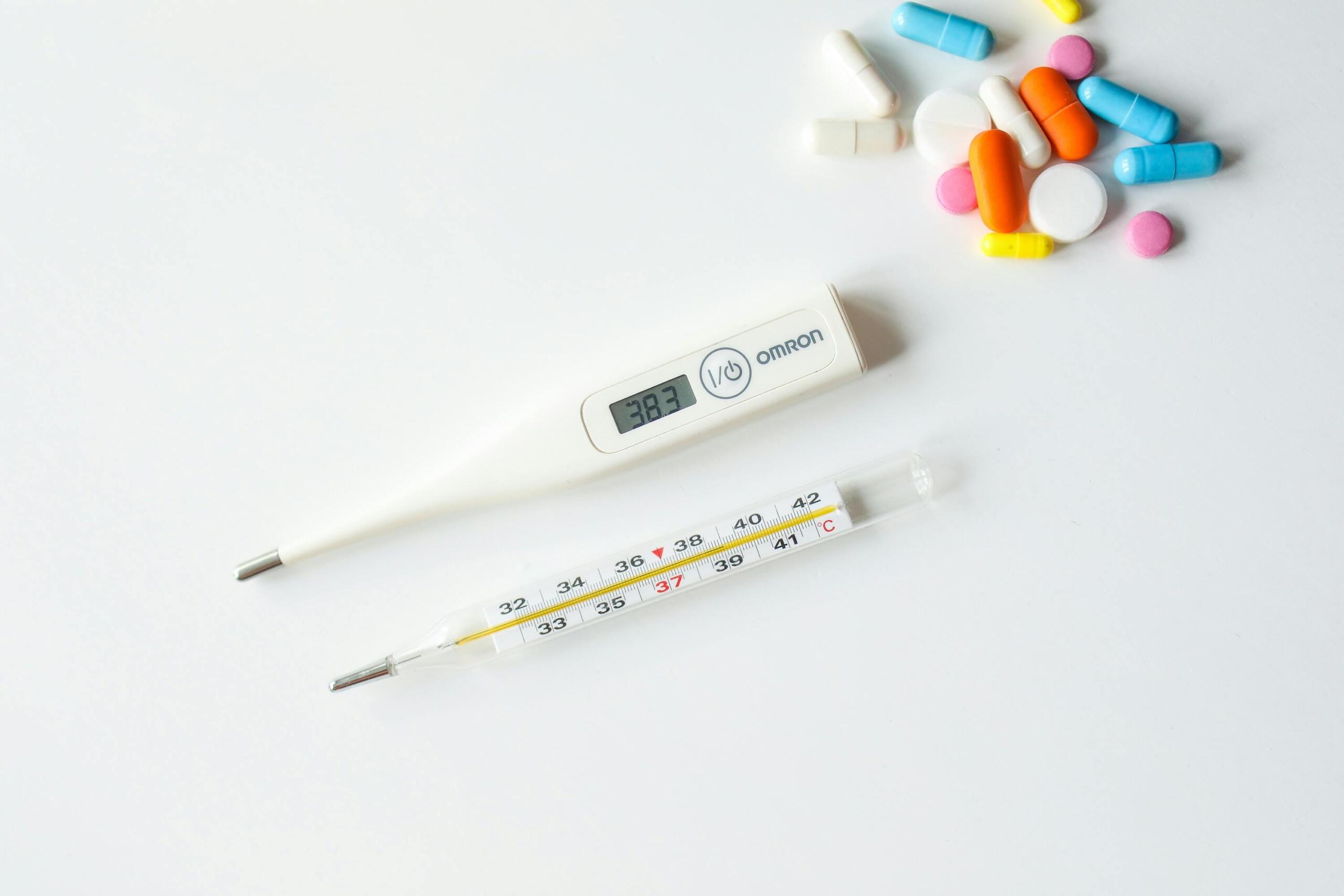Digitale en analoge thermometer naast een verzameling pillen, meting toont koorts van 38,7 °C.