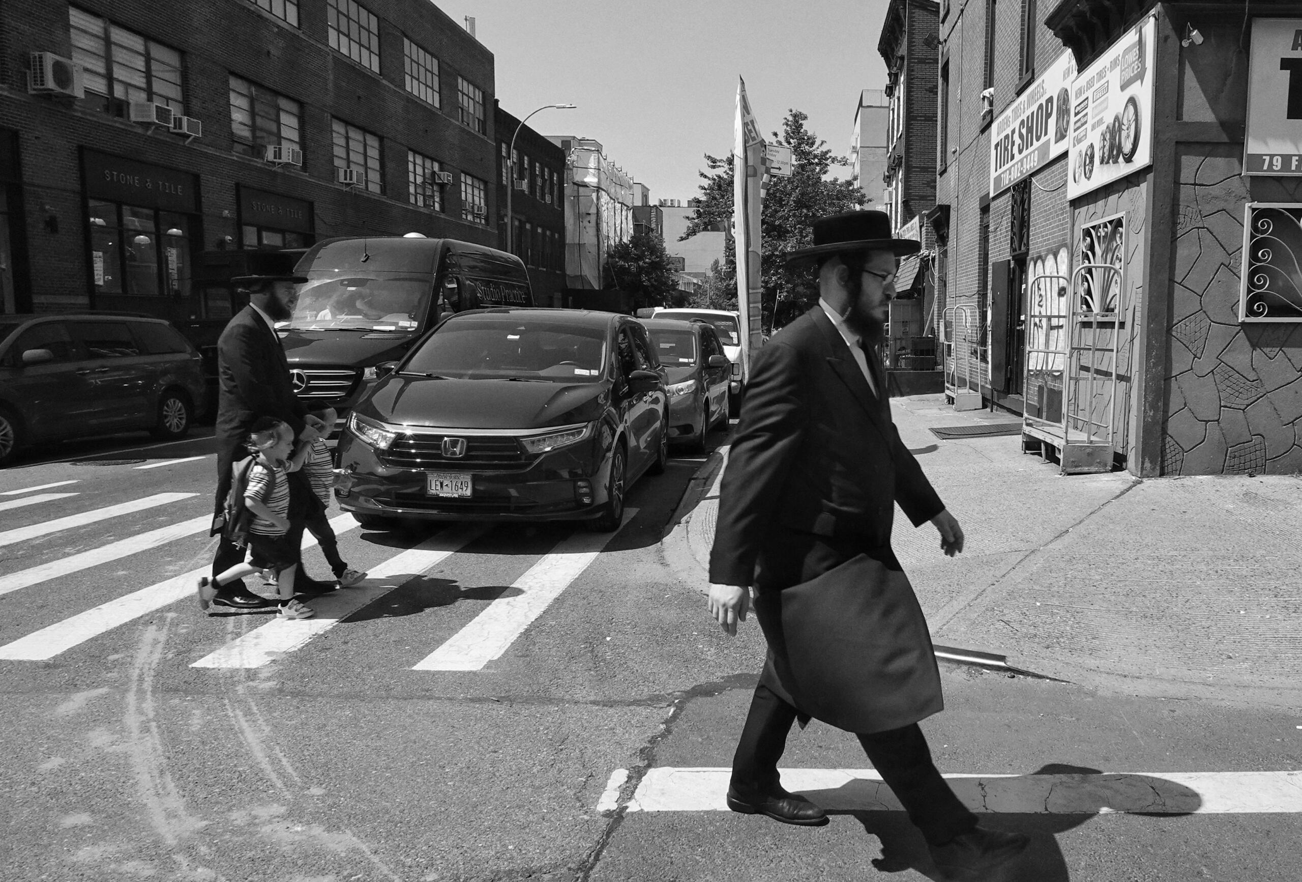 Orthodox-joodse mannen en kinderen steken een straat over in een stedelijke wijk, herkenbaar aan hun traditionele kleding.