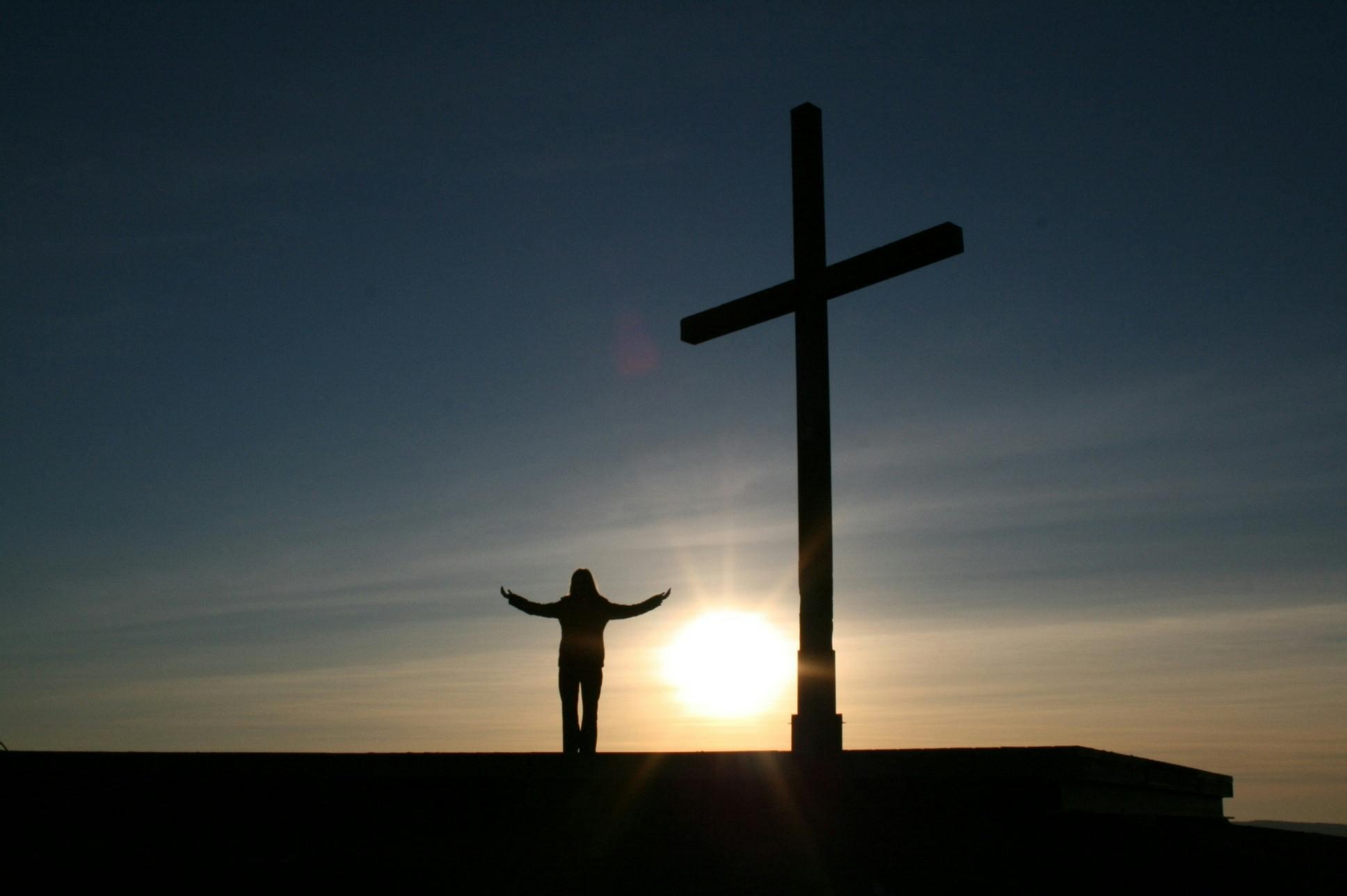 Een silhouet van een persoon bij een groot kruis bij zonsopkomst.