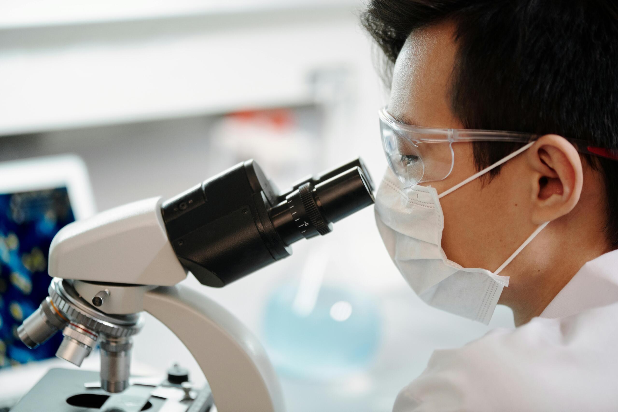Een man in witte laboratoriumjas zit in een moderne kantooromgeving achter een computer, met grafieken en tabellen zichtbaar op het scherm.
