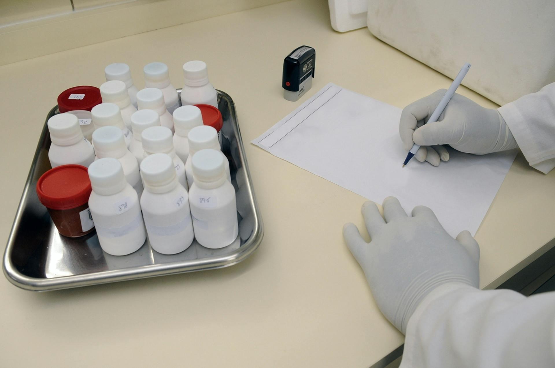 Een laborant in witte jas en handschoenen werkt in een laboratoriumomgeving en schrijft gegevens op, terwijl verschillende flesjes met witte doppen op een metalen dienblad staan.
