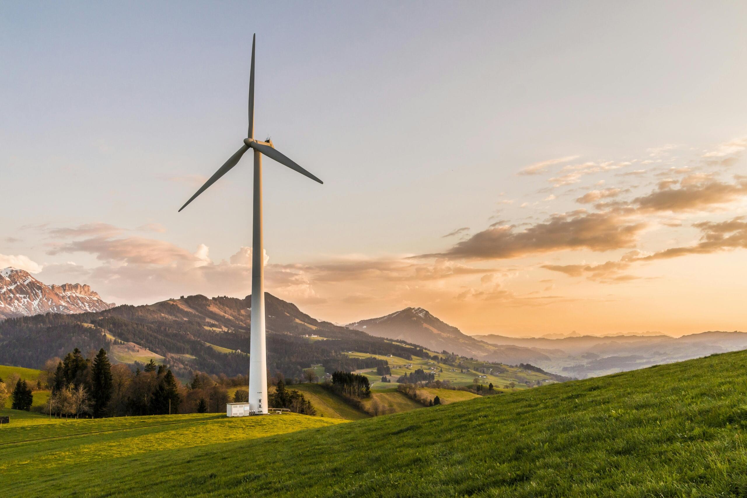 Een grote windturbine in een groen landschap met heuvels en bergen op de achtergrond, bij zonsondergang.