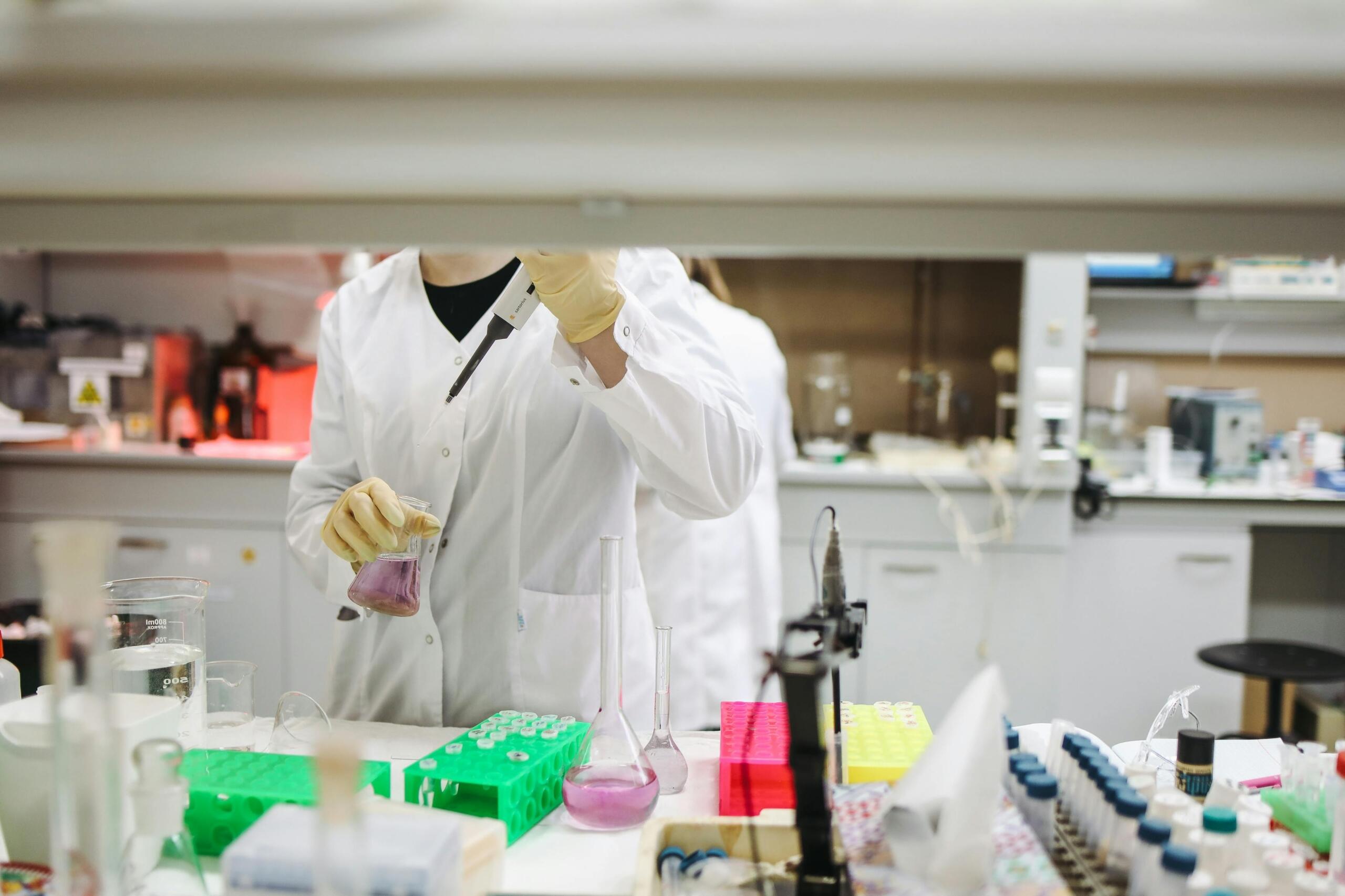 Een laborant in witte jas en gele handschoenen gebruikt een pipet en houdt een erlenmeyer met paarse vloeistof vast.