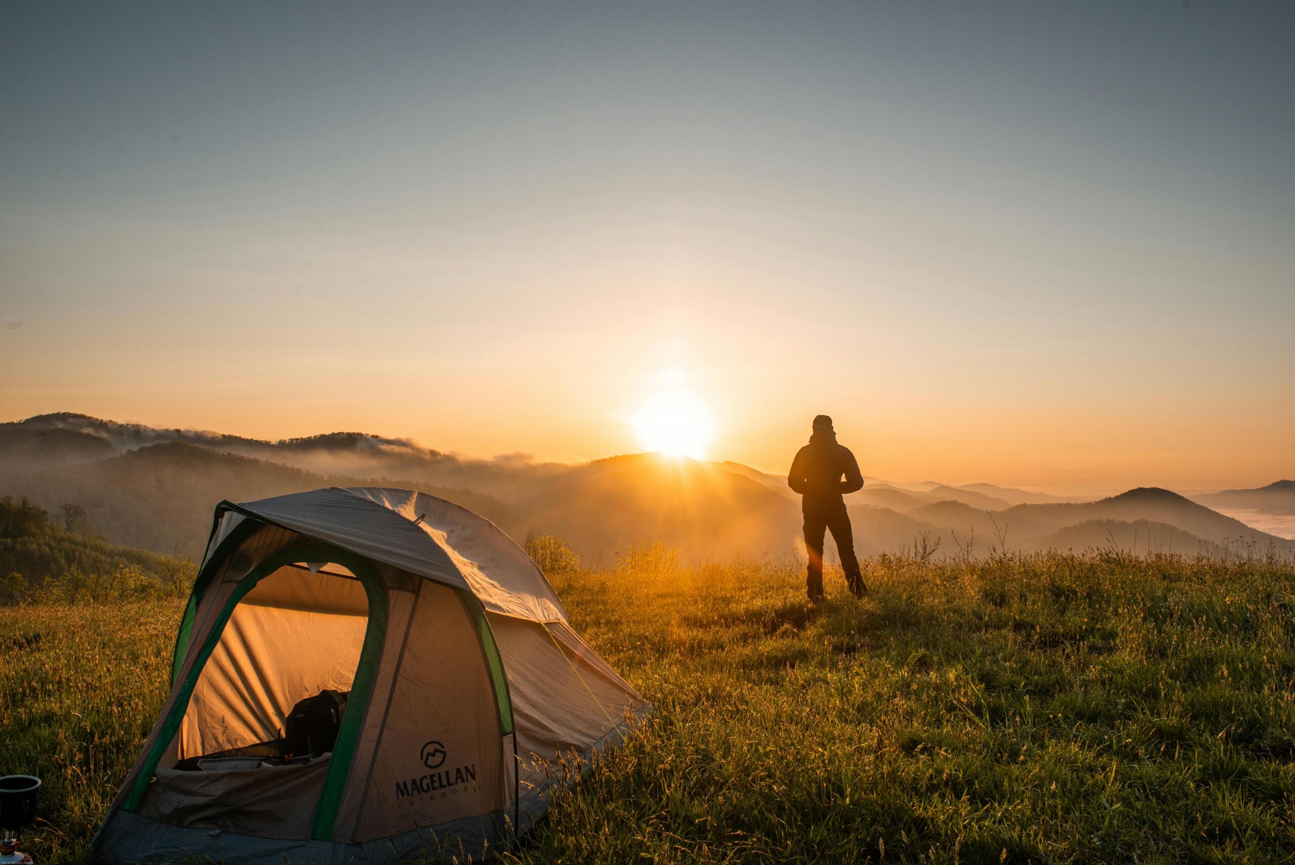 Een persoon staat naast een tent op een heuvel en kijkt naar de opkomende zon boven mistige bergen.