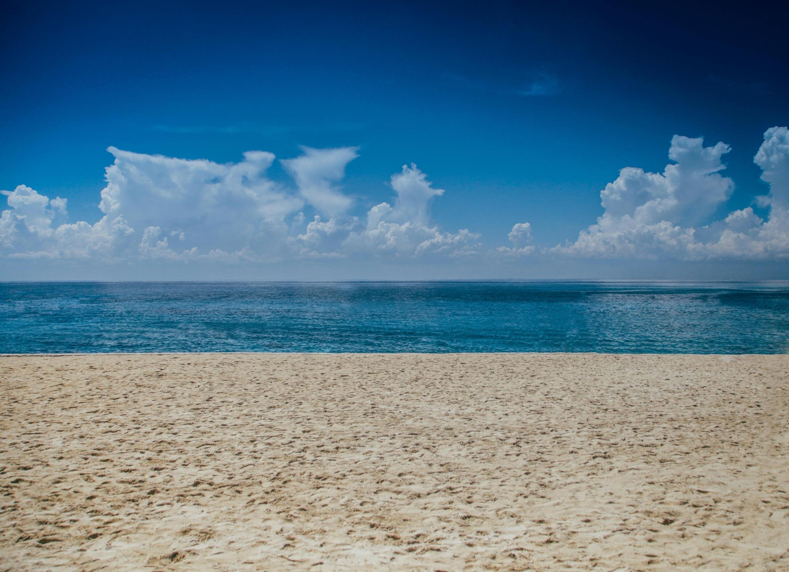 Een leeg, zonovergoten strand met helderblauw water en witte wolken aan de horizon.
