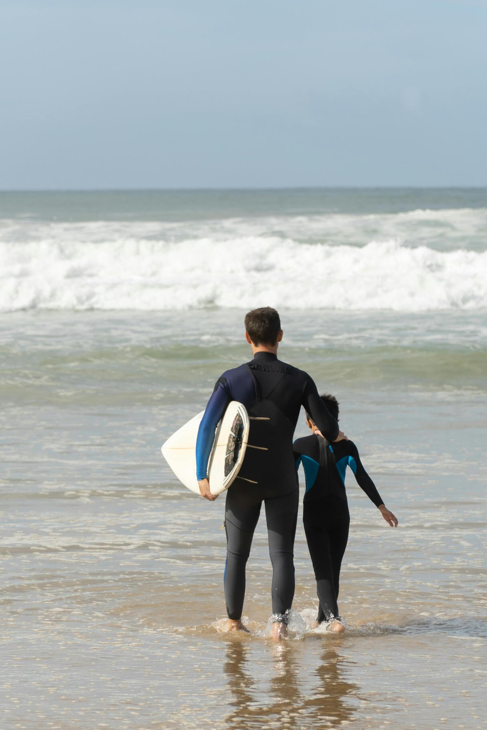 Een volwassene en een kind in wetsuits lopen met een surfplank het water in.