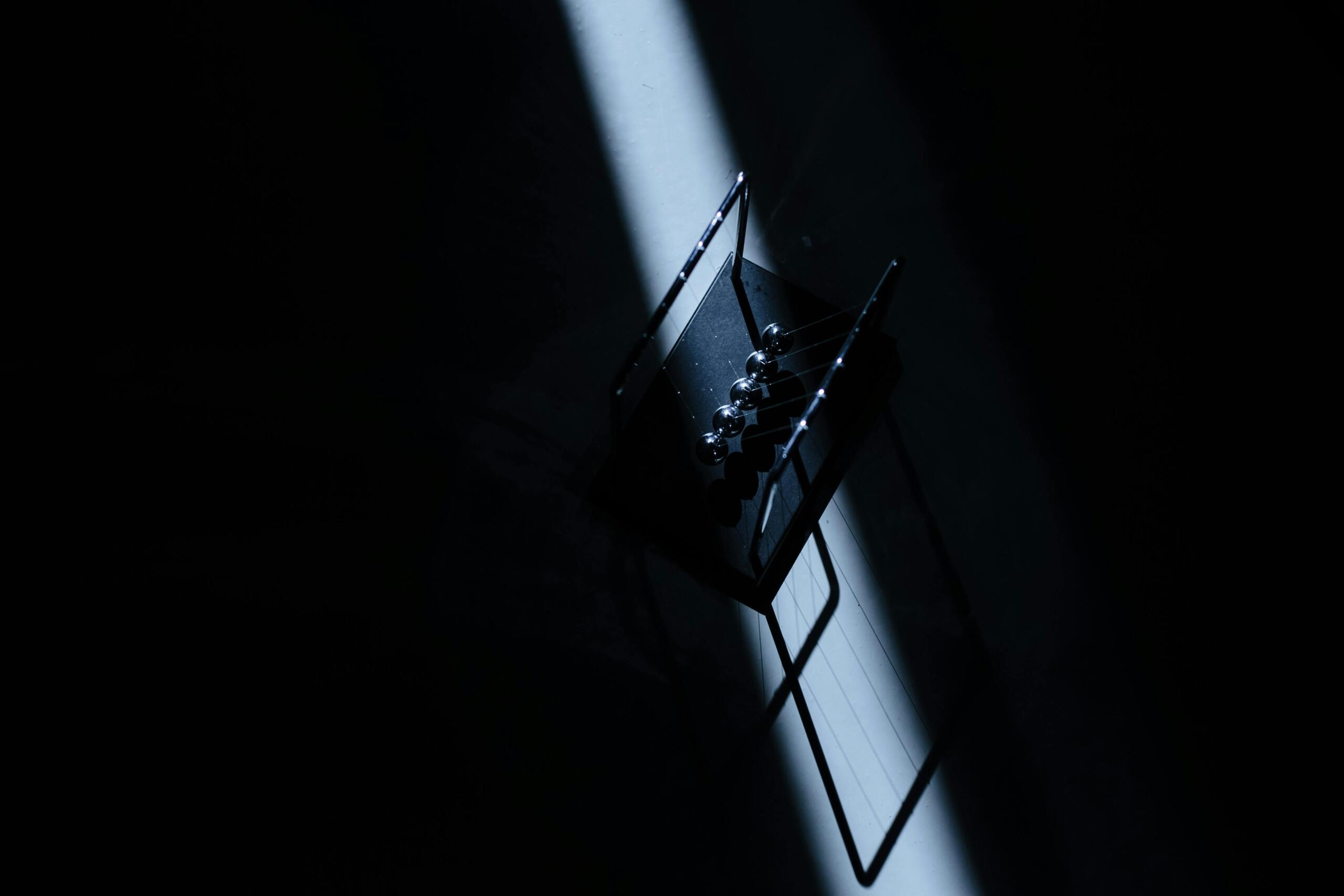 Een Newtonpendel wordt belicht door een smalle lichtstraal, waardoor de metalen kogels en hun schaduw scherp zichtbaar zijn in een donkere ruimte.