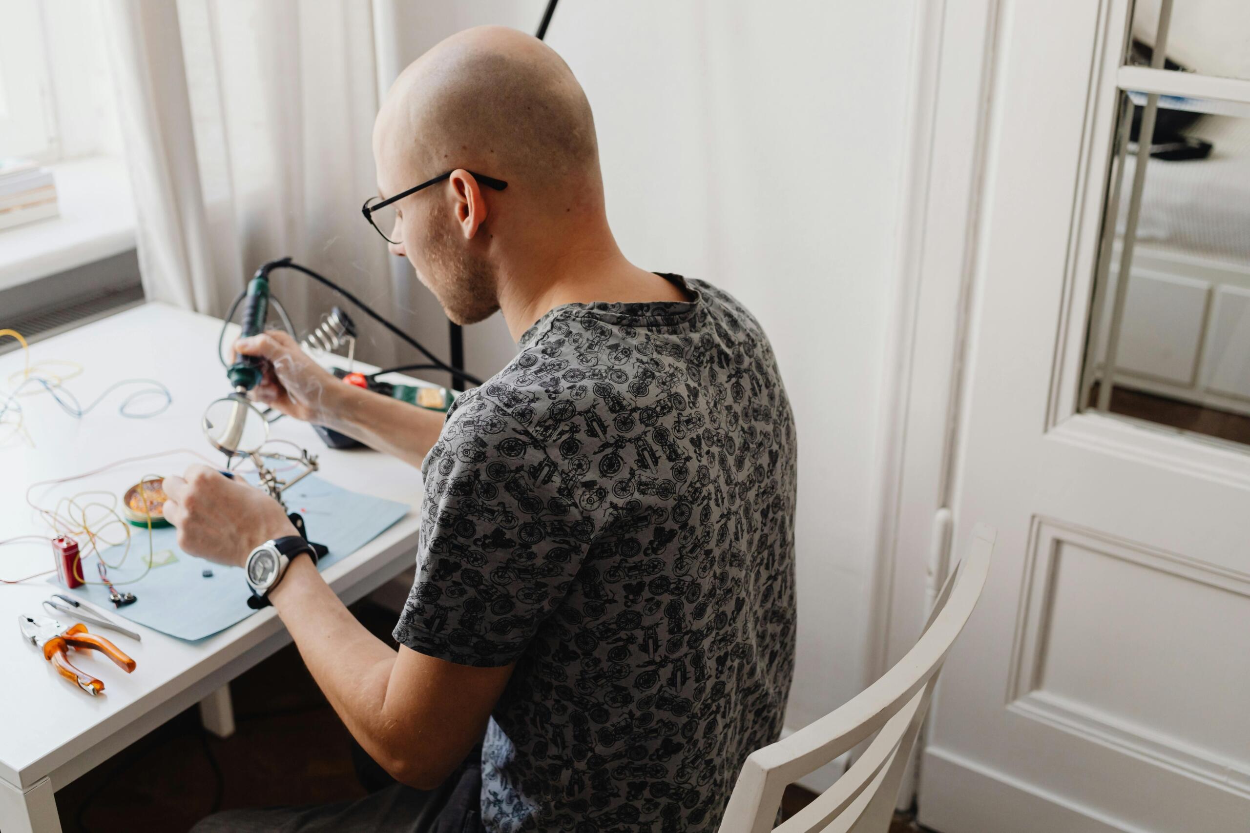 Een man met een bril soldeert elektronische onderdelen aan een witte tafel met gereedschap en draden.