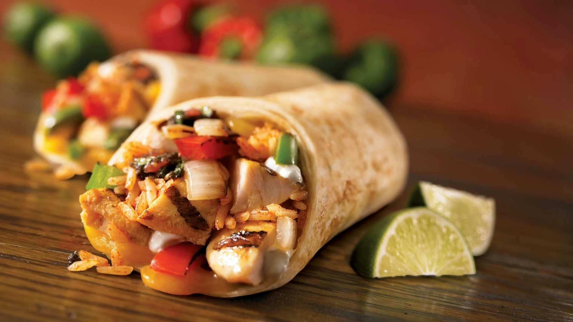 Close‑up van een tortilla wrap gevuld met gegrilde kip, groenten en rijst op een houten oppervlak met partjes limoen ernaast.