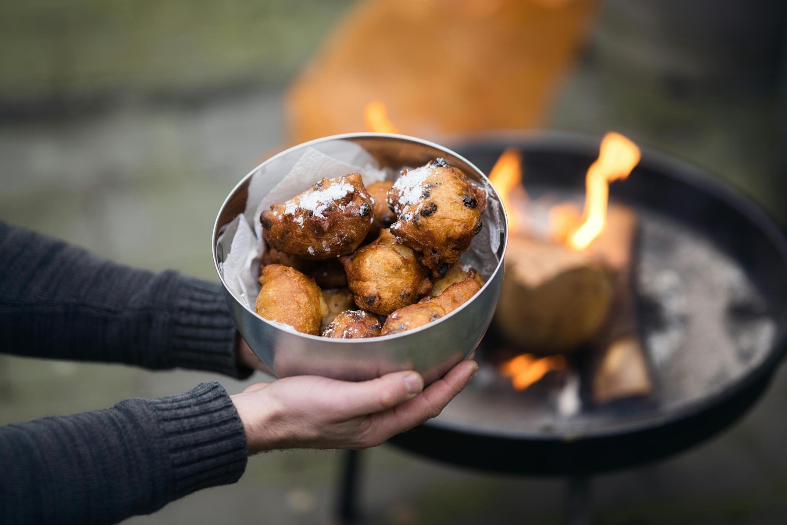 Een persoon in een donkere trui houdt een metalen kom met goudbruine oliebollen vast; de oliebollen zijn bestrooid met poedersuiker en in de achtergrond is een buitenvuur te zien