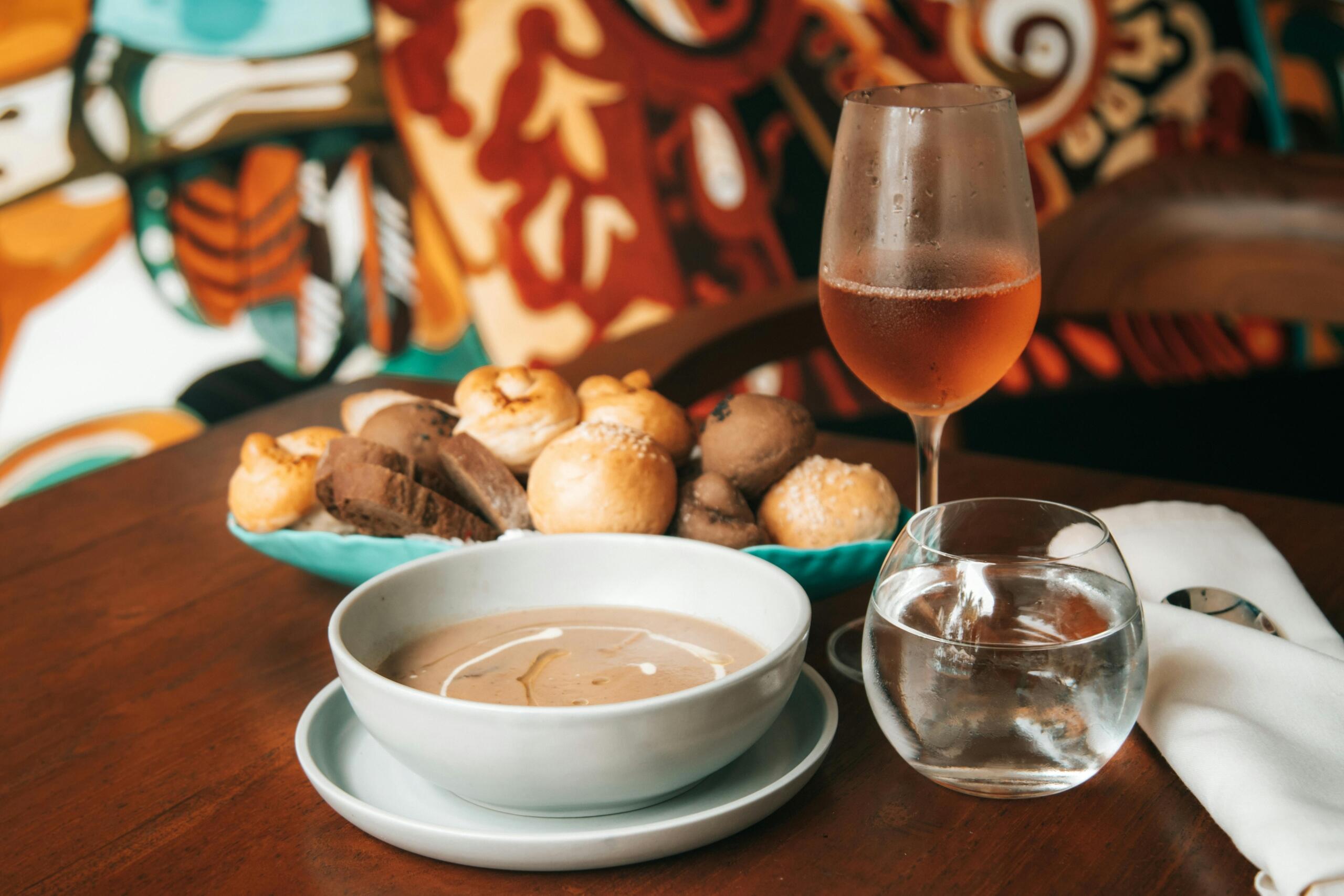 Op een houten tafel staan een grijze kom met romige soep met een swirl van room, een schaal met verschillende broodjes en plakken donker brood, een waterglas en een glas rosékleurige wijn.