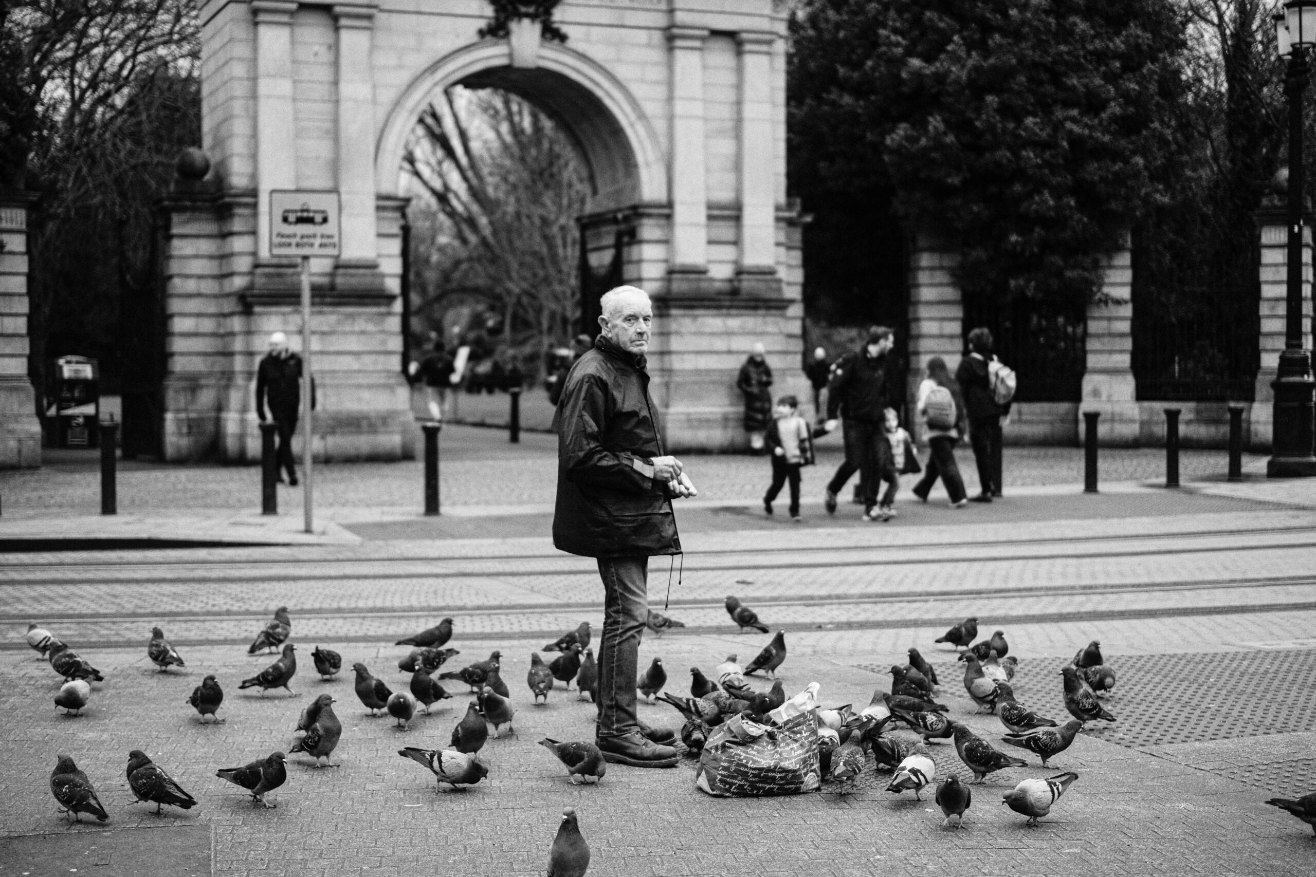 Een oudere man staat tussen duiven op een druk plein in Dublin, met een stenen boogpoort op de achtergrond.