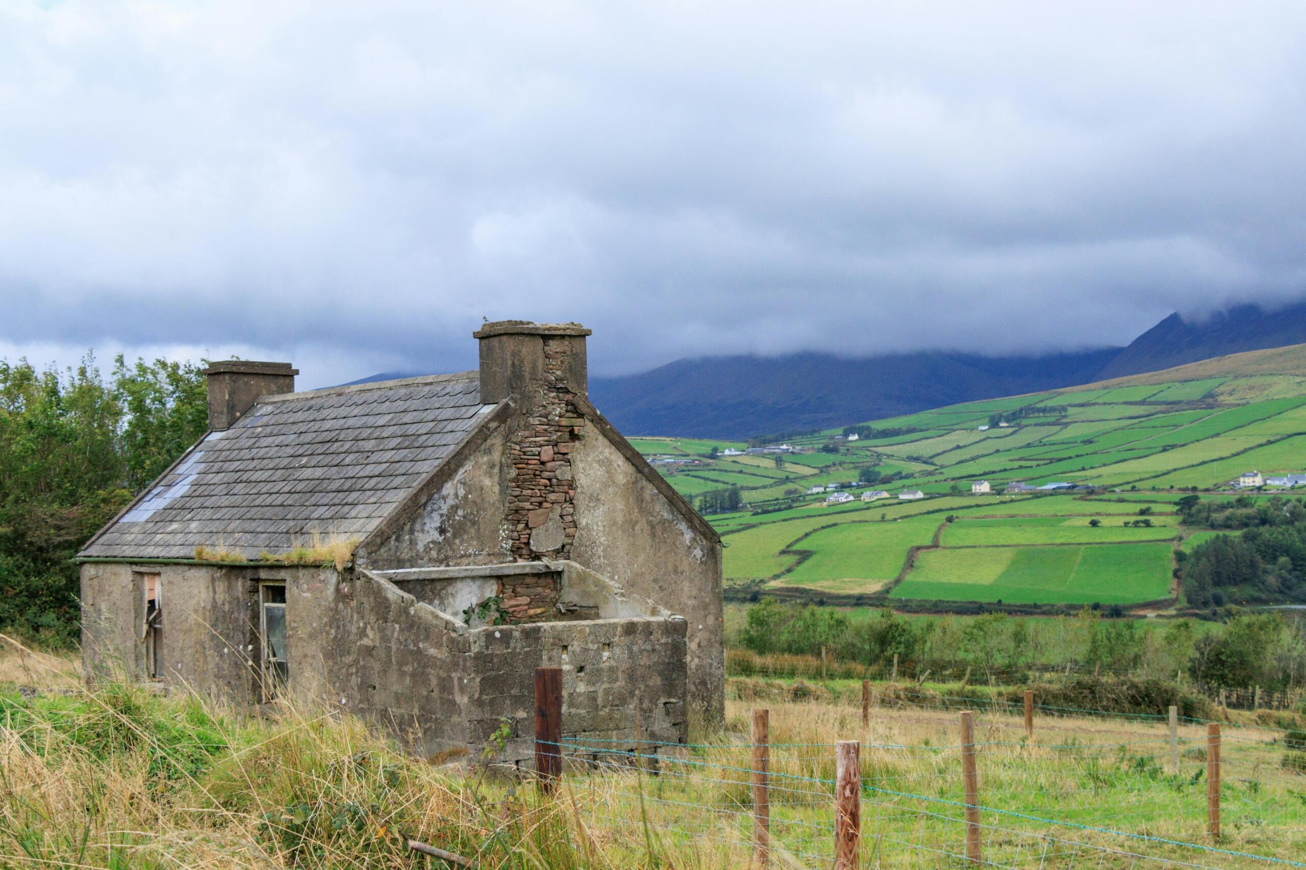 Een oud stenen huisje tussen groene lappendekenvelden, met rollende heuvels en laaghangende wolken op de achtergrond.