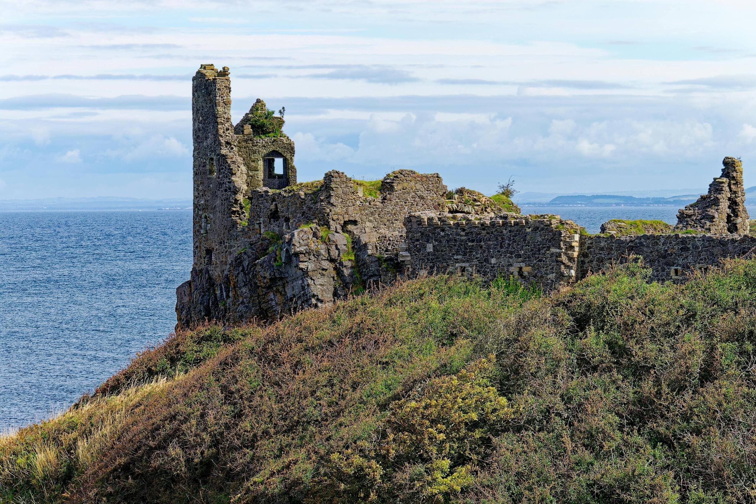De ruïnes van een oude stenen structuur op een kustklif, omringd door ruige begroeiing en uitkijkend over de zee.