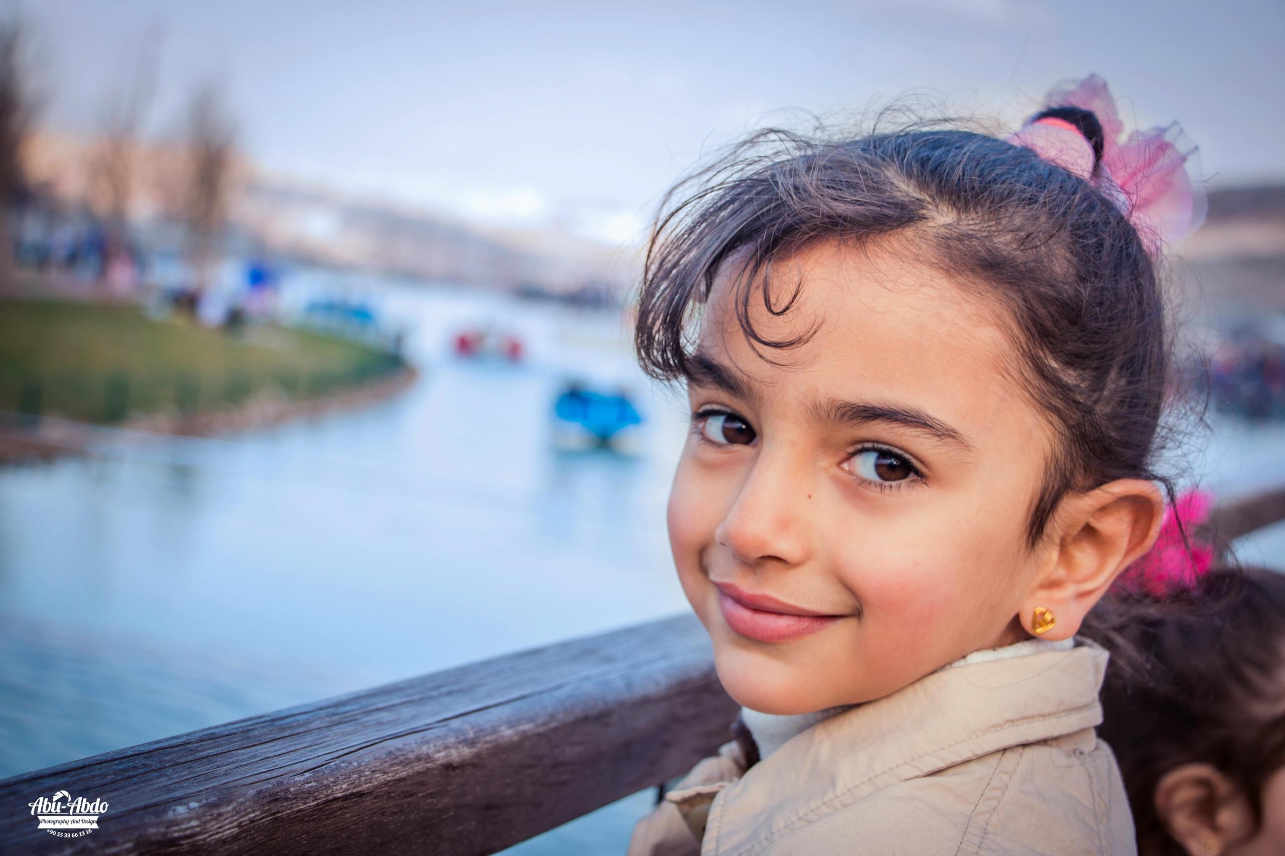 Een meisje kijkt glimlachend over haar schouder terwijl ze leunt op een houten railing bij een rustige waterkant.