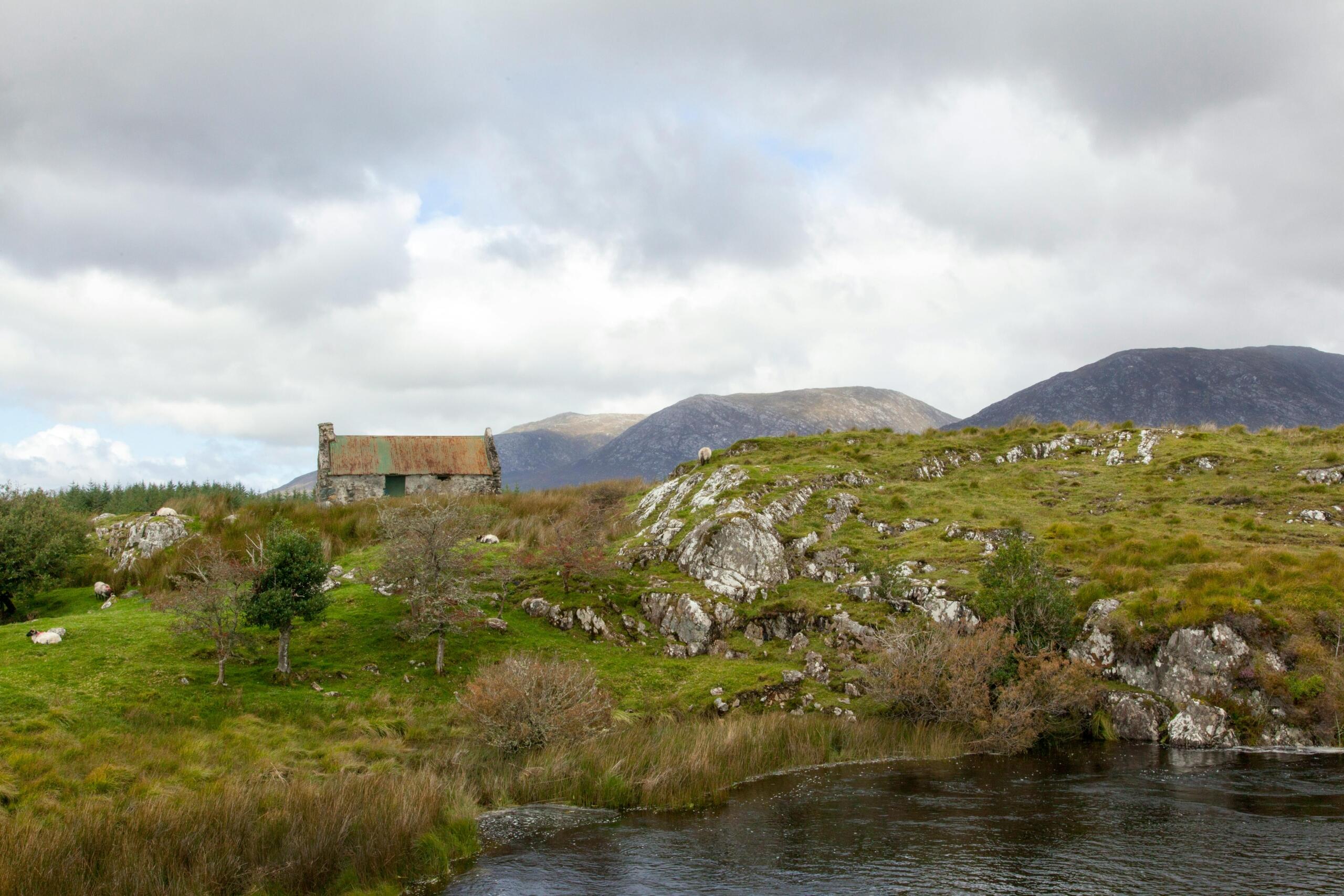 Een ruwe stenen hut staat tussen rotsen, graslanden en een rivier, met bergen op de achtergrond.