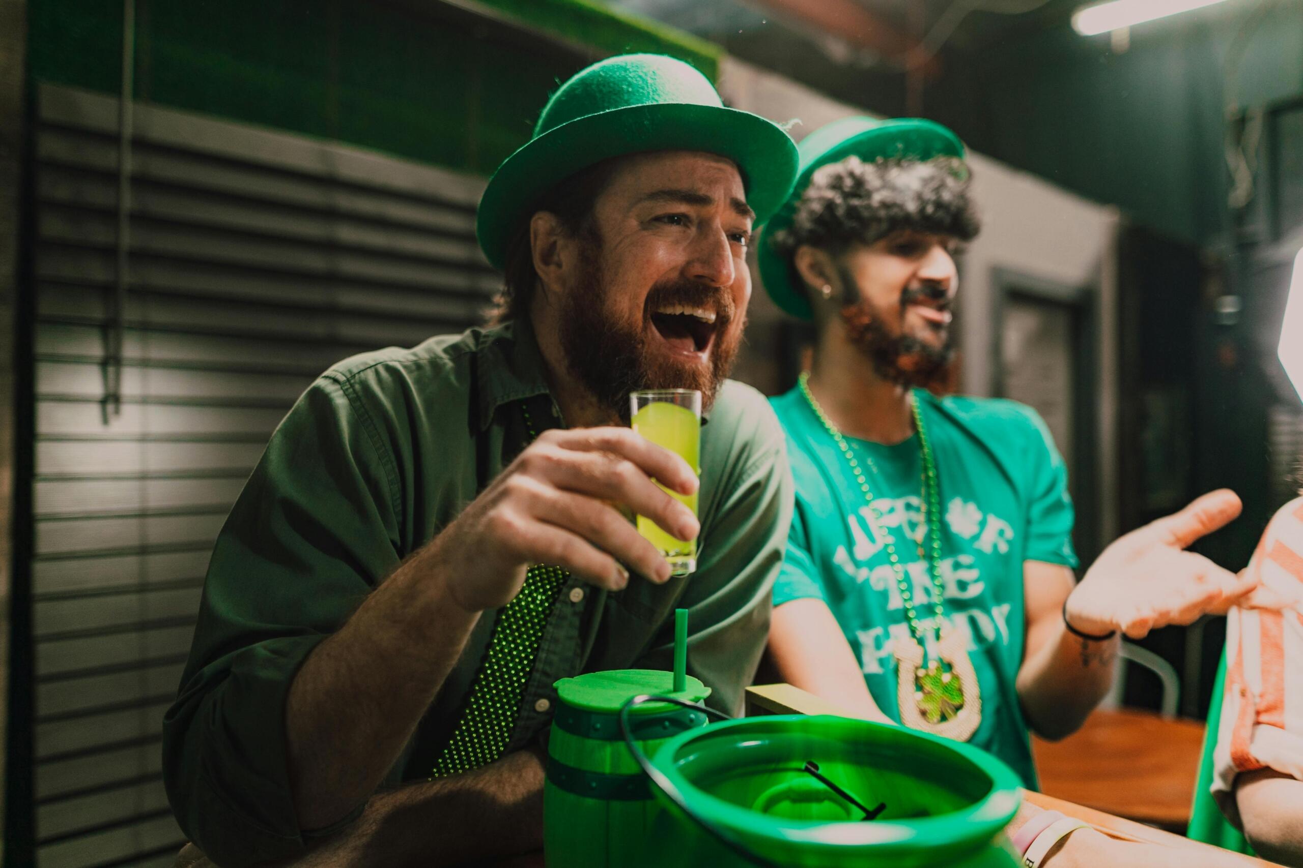 Twee mannen in groene kleding en hoeden zitten aan de bar en lachen terwijl een van hen een glas met groene drank vasthoudt.