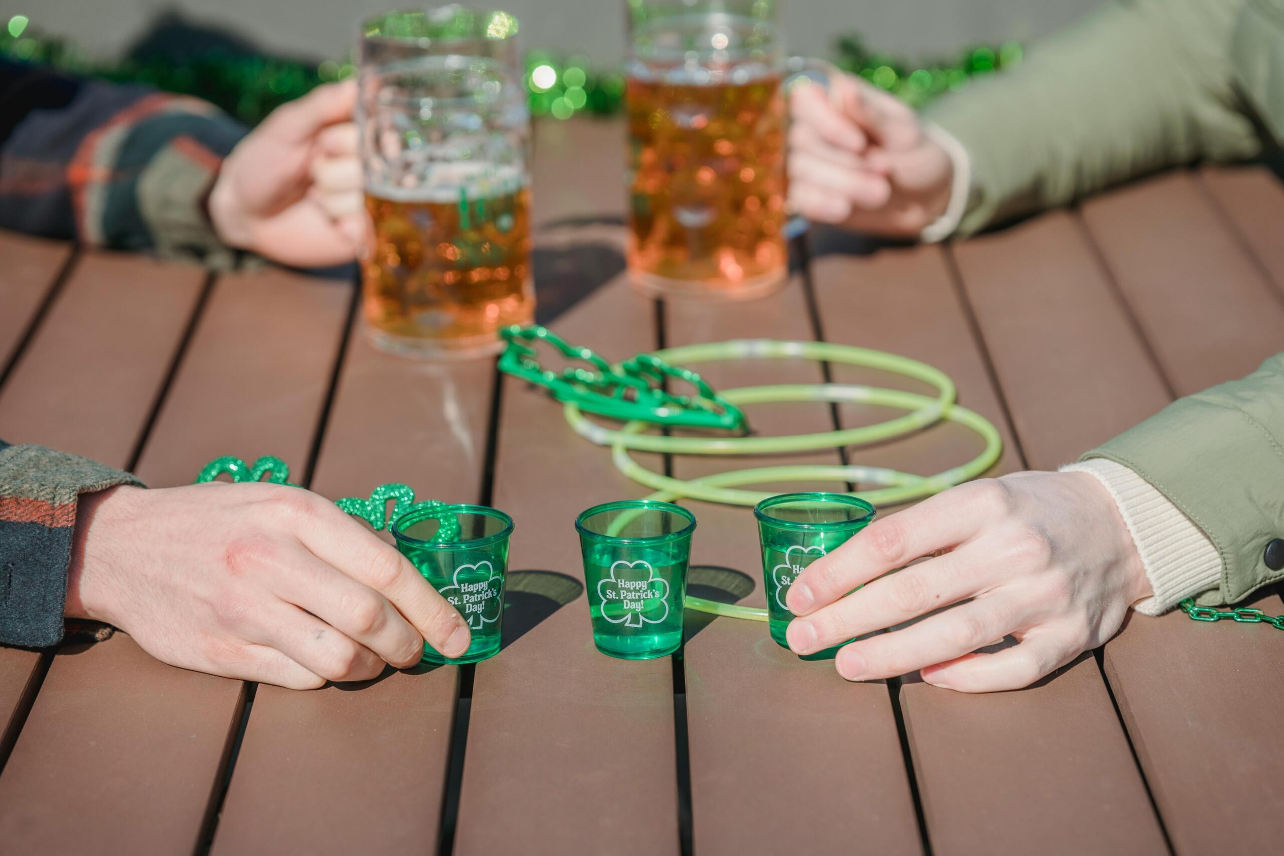 Drie groene shotglaasjes worden vastgehouden door verschillende handen, terwijl op de achtergrond glazen bier en groene feestaccessoires op de tafel liggen.