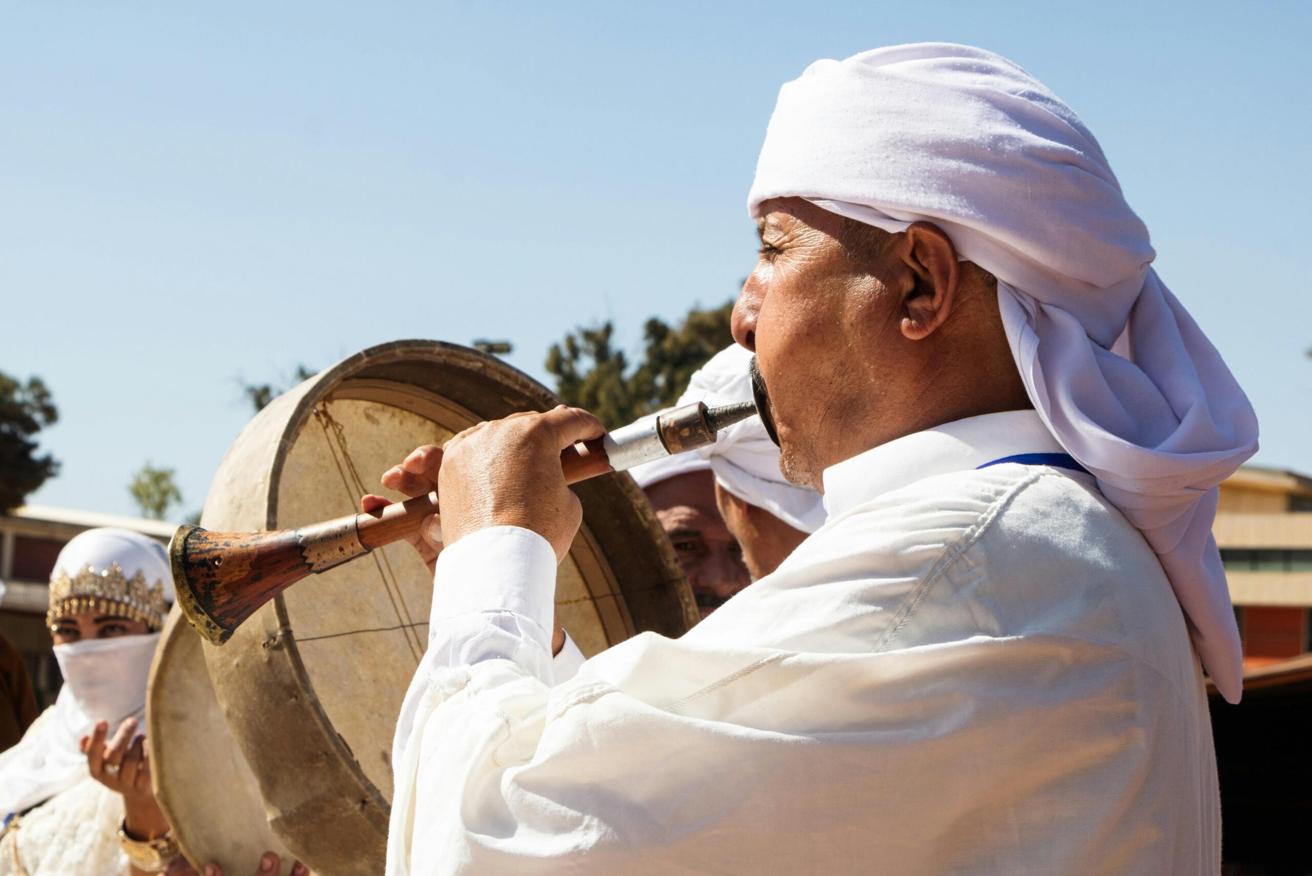 Een man met een witte hoofddoek speelt op een zurna, een dubbelrietinstrument, terwijl een andere persoon op een grote trom slaat. Op de achtergrond staat een gesluierde vrouw met een rijk versierde hoofdtooi.