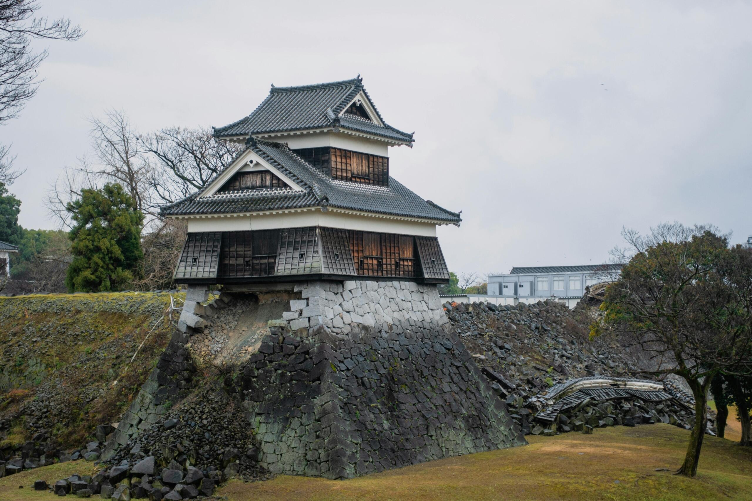Kleine houten toren van het kasteel van Kumamoto op een beschadigde stenen basis, met ingestorte muren en puin eromheen, kale bomen en een bewolkte lucht op de achtergrond.