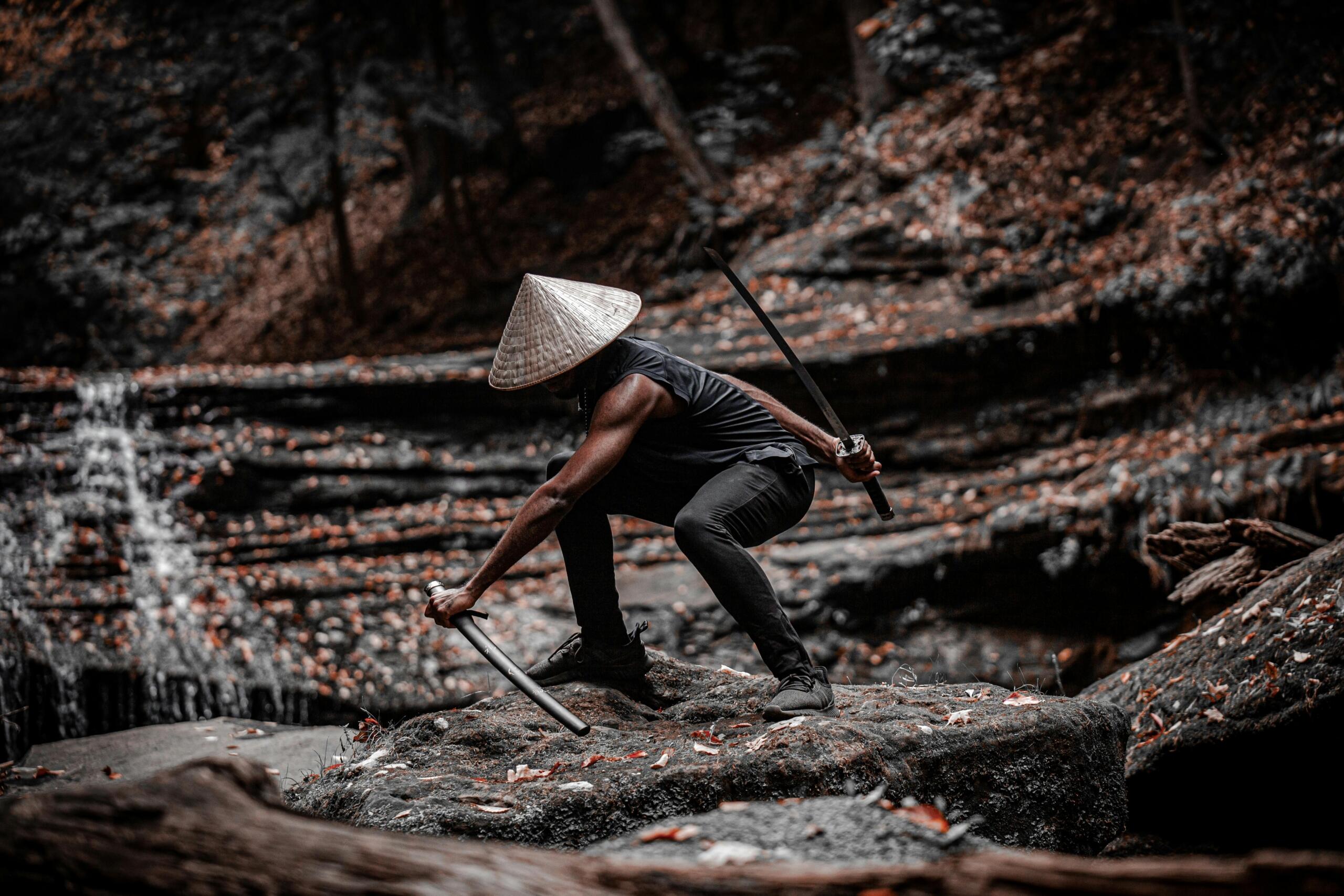 Man met een kegelvormige strohoed die gehurkt op een met mos bedekte rots zit in een bos, twee zwaarden stevig vasthoudend, met een wazige beek en herfstbladeren op de achtergrond.