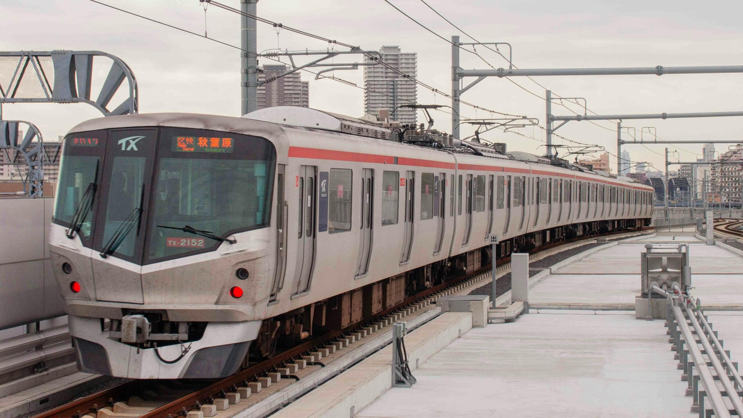 Moderne zilverkleurige forenzentrein die over verhoogde spoorrails rijdt, met bovenleidingen en stadsskyline zichtbaar op de achtergrond.