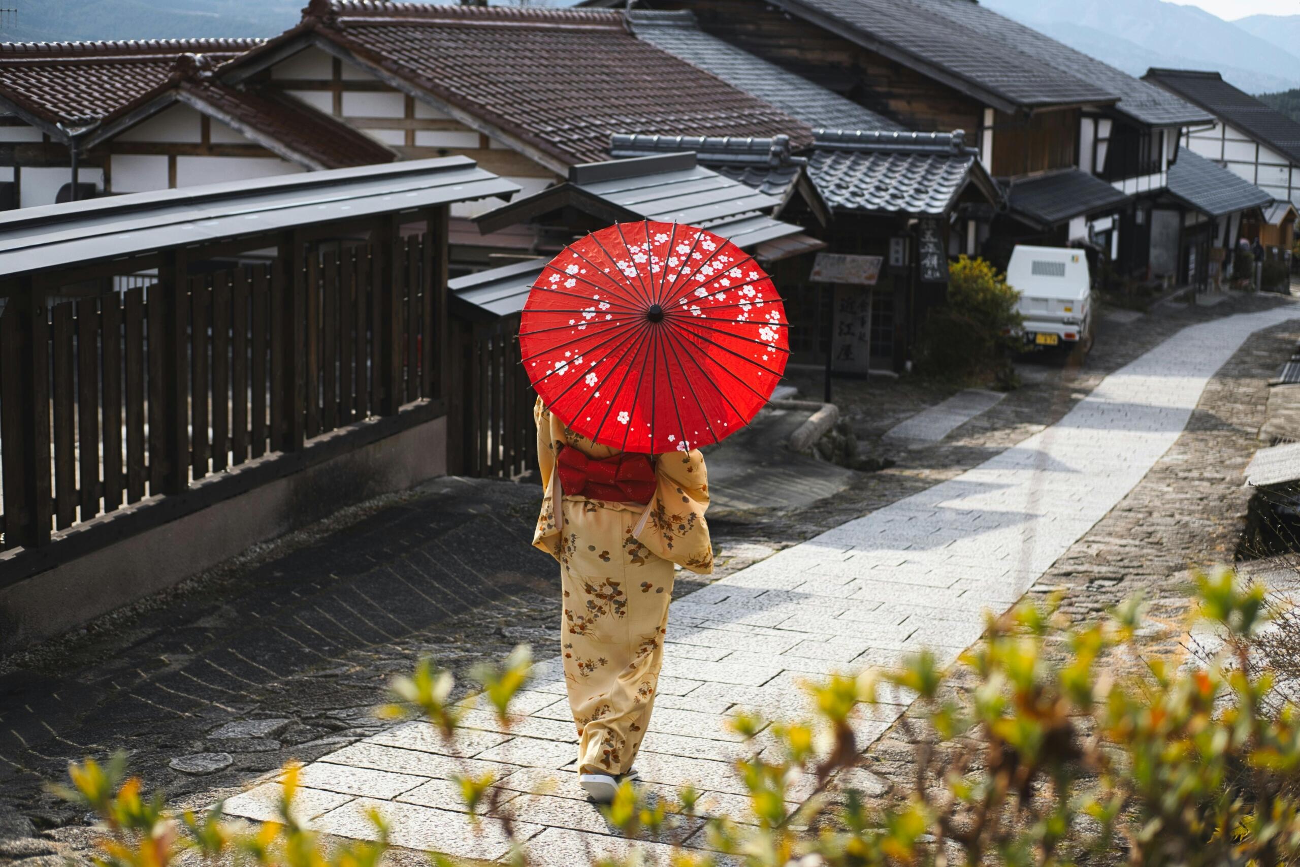 Achteraanzicht van een vrouw in een kimono met patroon die een rode parasol draagt terwijl zij over een met stenen geplaveide straat loopt, omzoomd door traditionele houten huizen.