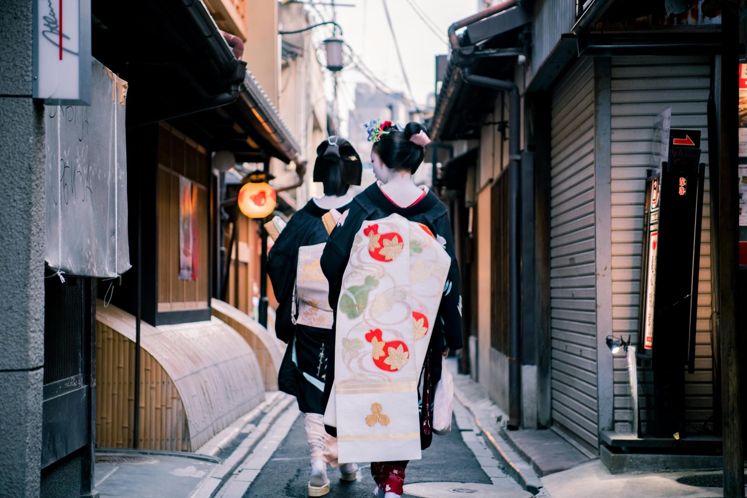 Twee vrouwen in traditionele kimono lopen door een smalle straat in Kyoto, met houten gebouwen en lampionnen aan weerszijden.