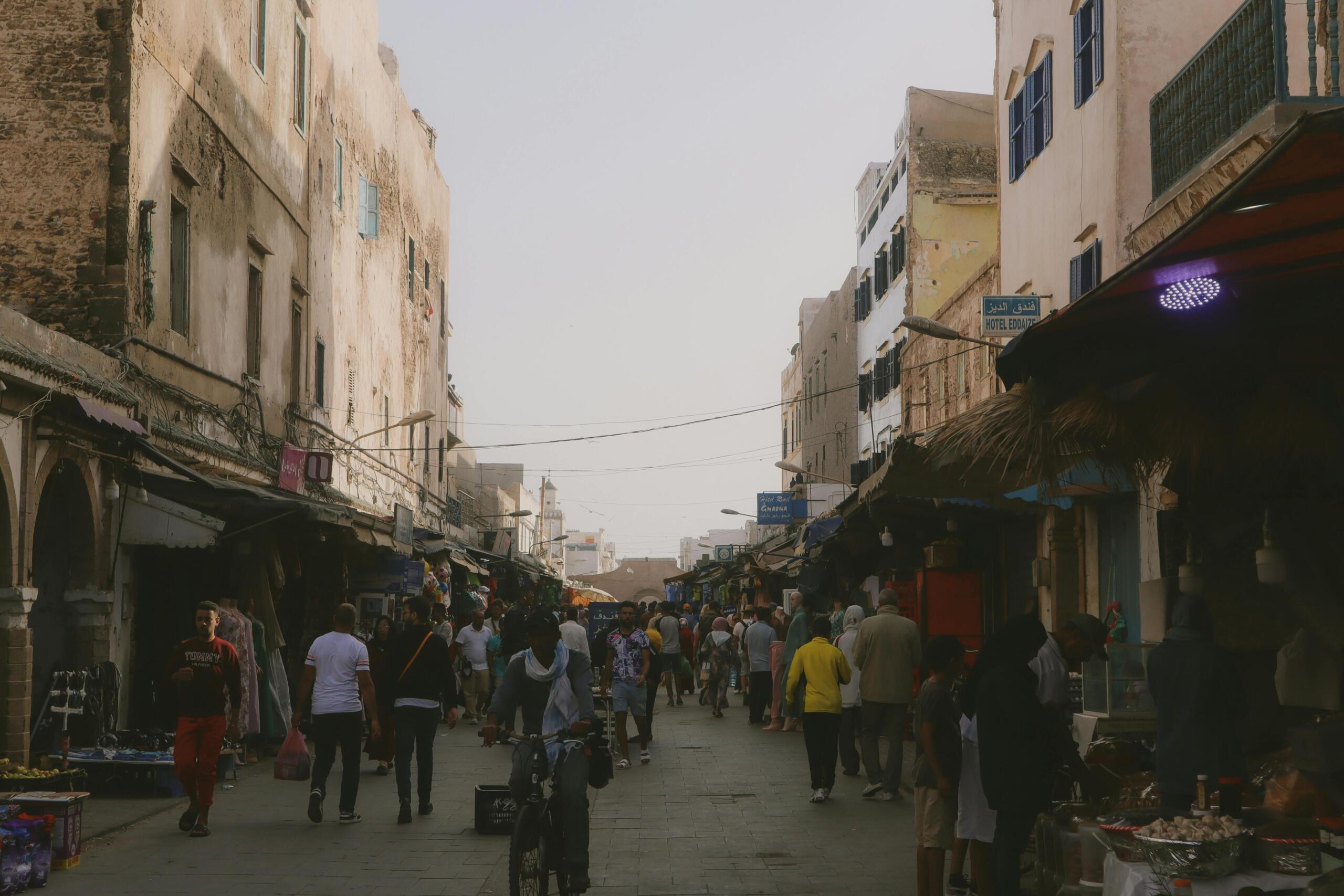 Levendige scène van een historische marktplaats met voetgangers die tussen oude gebouwen en kraampjes lopen in een smalle straat vol verkopers.