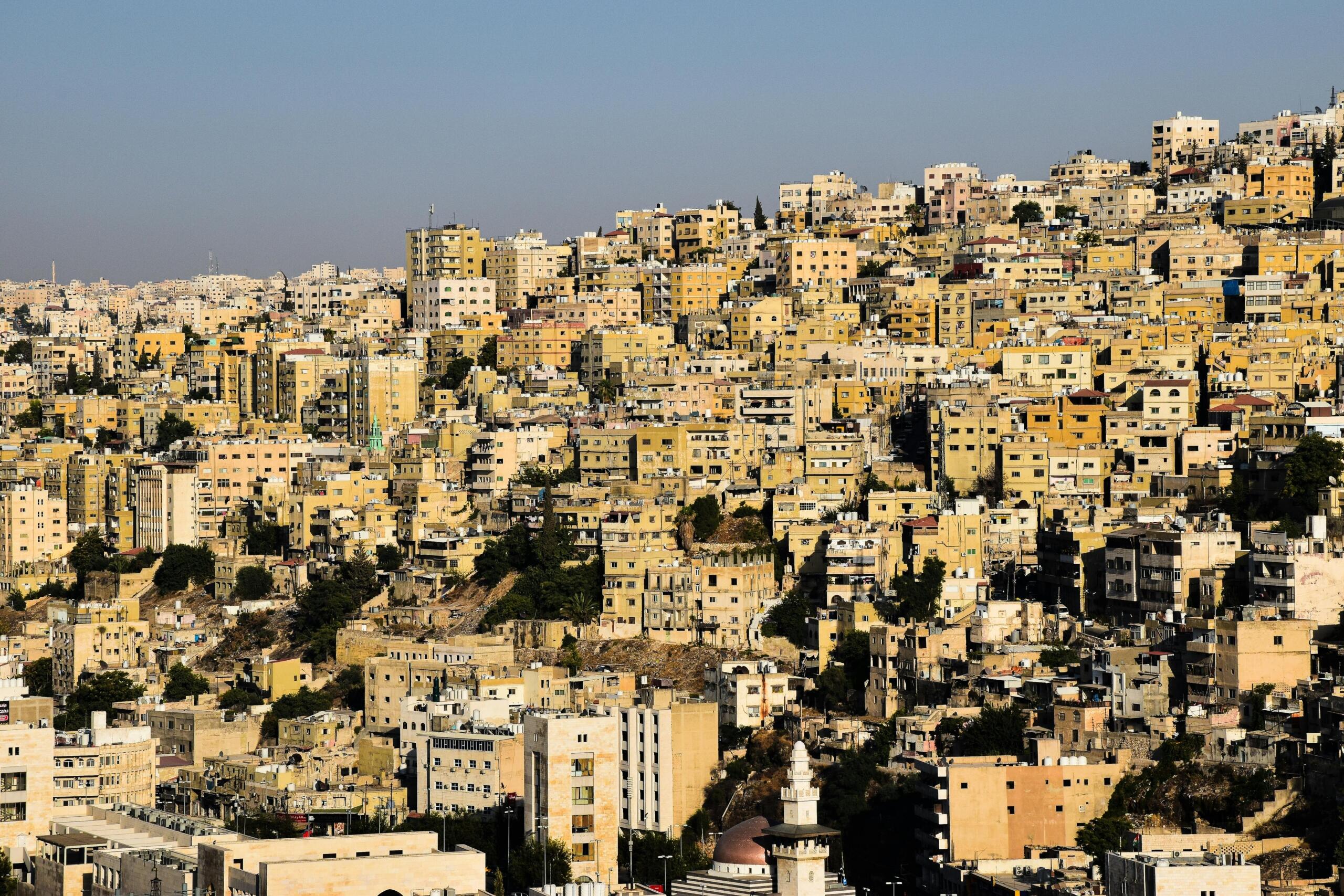 Uitzicht op een dichtbebouwd heuvelachtig stadslandschap in Amman, Jordanië, met beige en gele gebouwen die dicht op elkaar zijn gestapeld onder een heldere blauwe lucht.