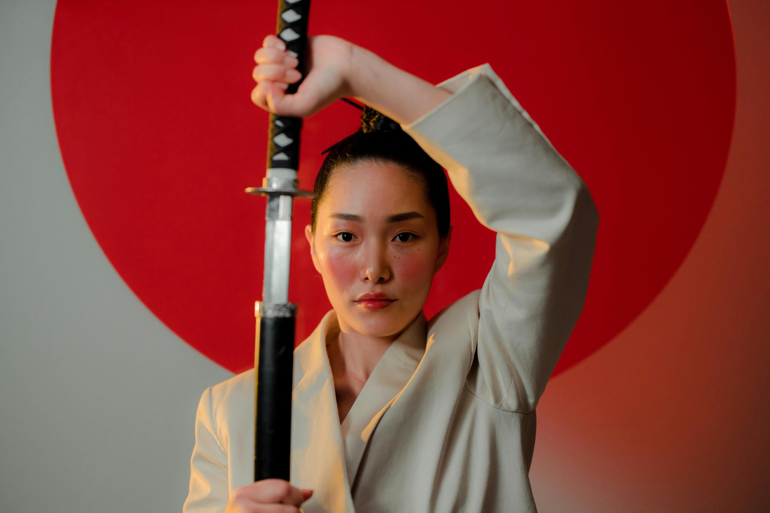 Vrouw in een wit jasje houdt een katana verticaal omhoog met geheven arm tegen een opvallende rode cirkel op de achtergrond.