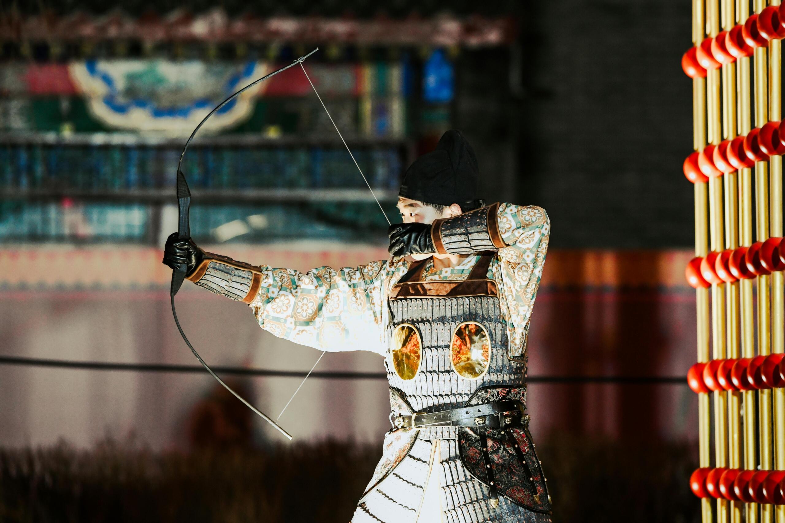 Man in traditioneel Japans harnas richt een boog en pijl tijdens een buitendemonstratie, met mensen en houten gebouwen op de achtergrond.