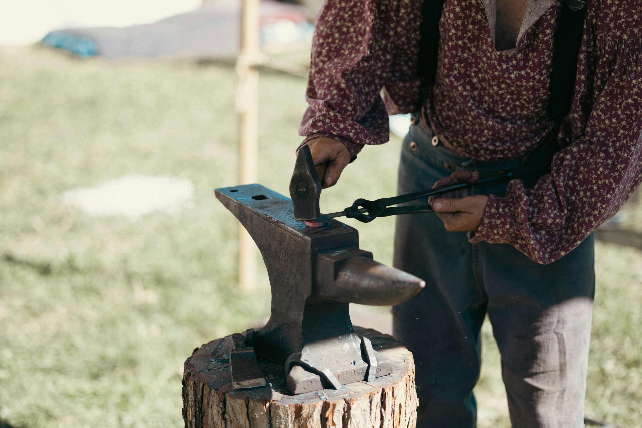 Smid in traditionele kleding slaat buiten op heet metaal op een aambeeld, met rook rond de smidse en houten bouwwerken in de buurt.