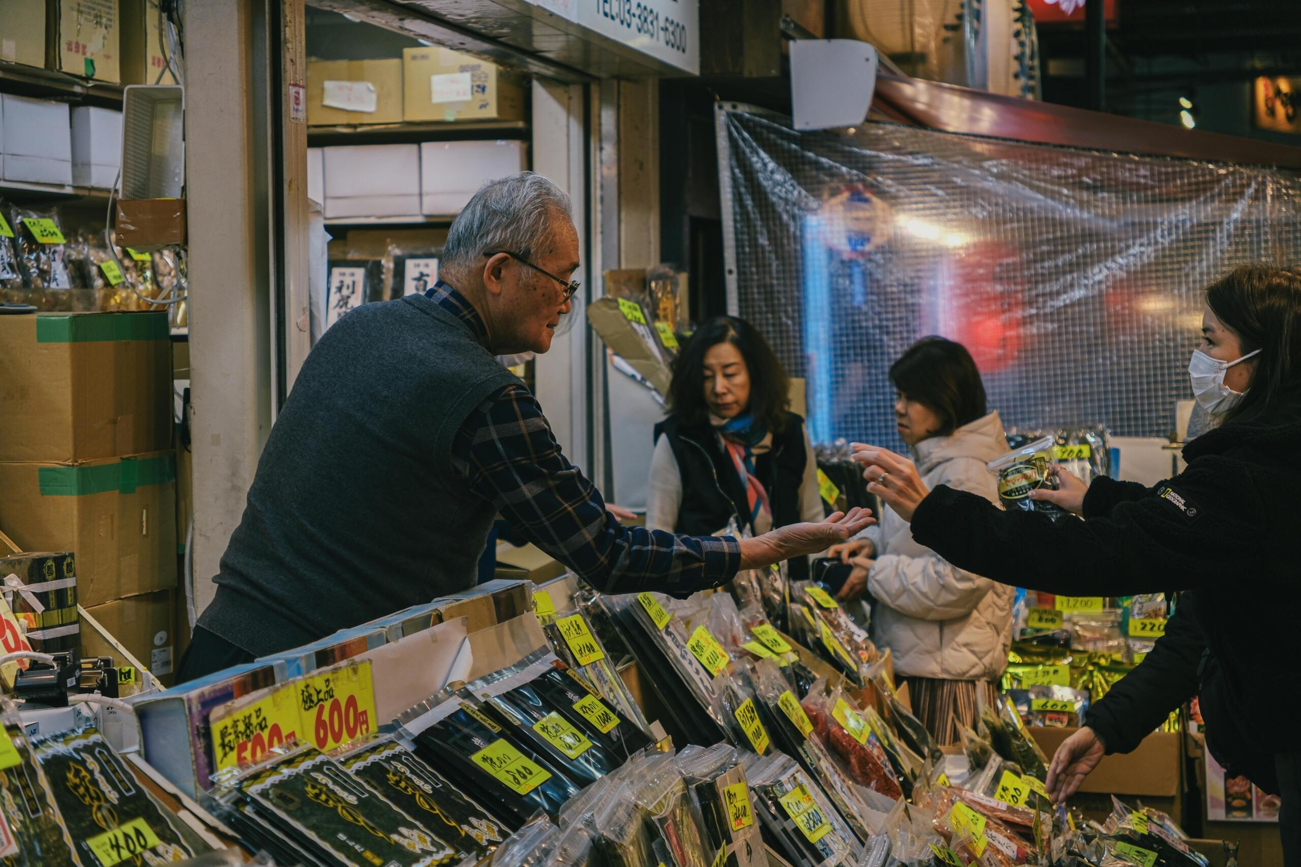Drukke Japanse straatmarkt waar een verkoper producten overhandigt aan klanten die mondkapjes dragen, omringd door manden met verse etenswaren.