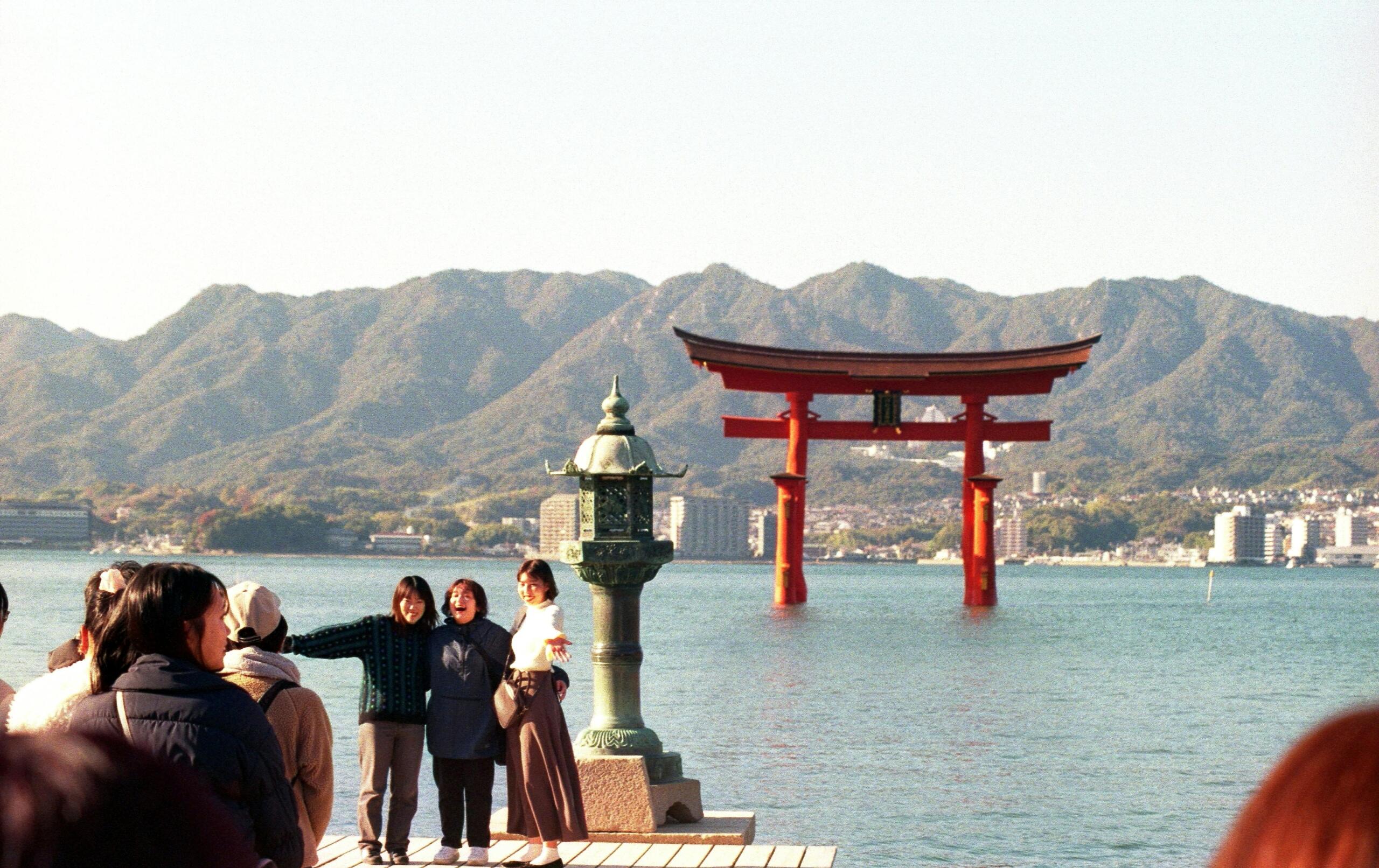 Rode torii-poort die in het water staat bij het Itsukushima-heiligdom op Miyajima, met bergen en een stad op de achtergrond, terwijl toeristen vanaf een pier toekijken.