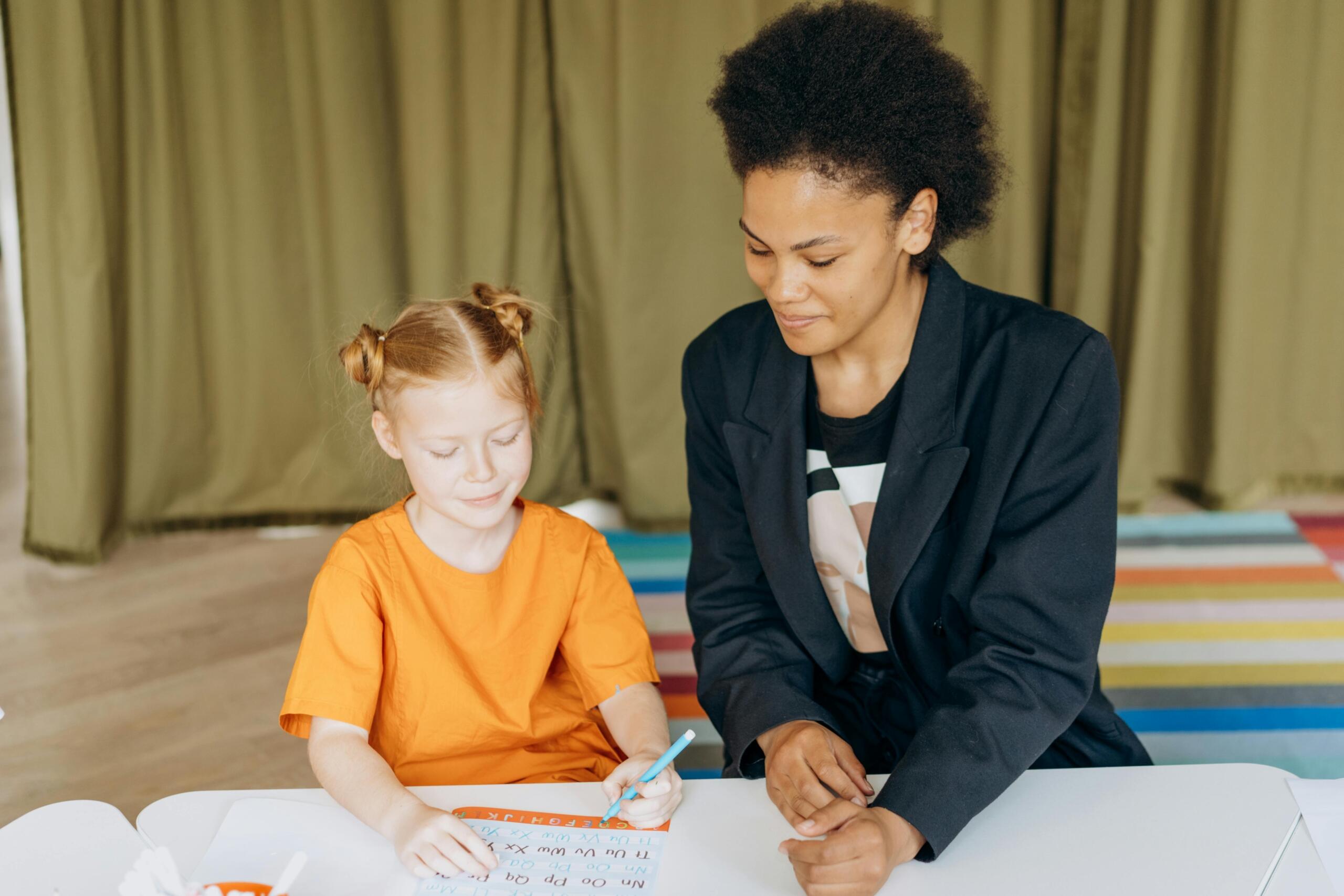 Vrouw met donker jasje zit naast een meisje met oranje trui aan een tafel en kijkt lachend naar haar werk.