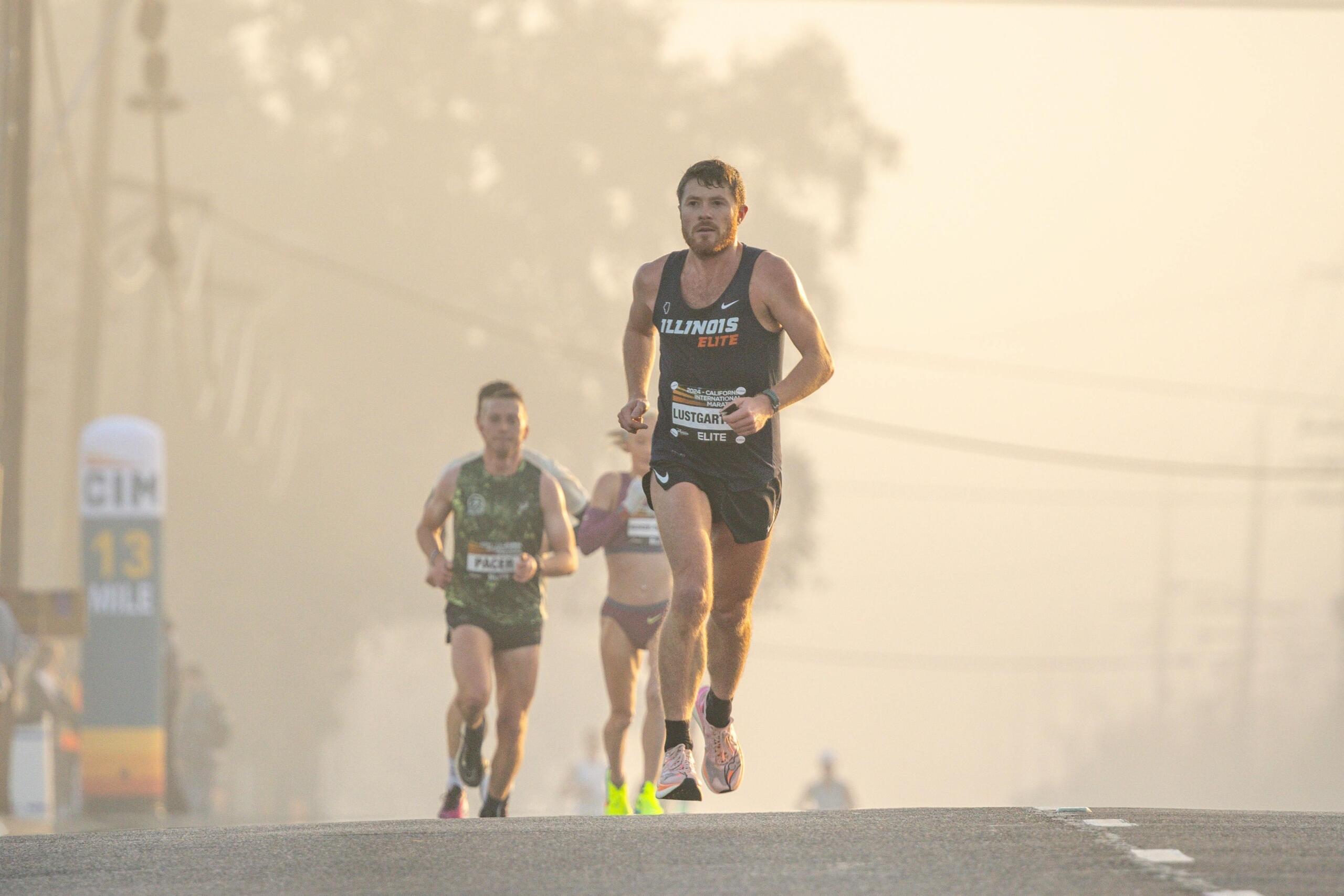 Marathonloper rent voorop op een brede weg met twee lopers die hem volgen in ochtendmist.