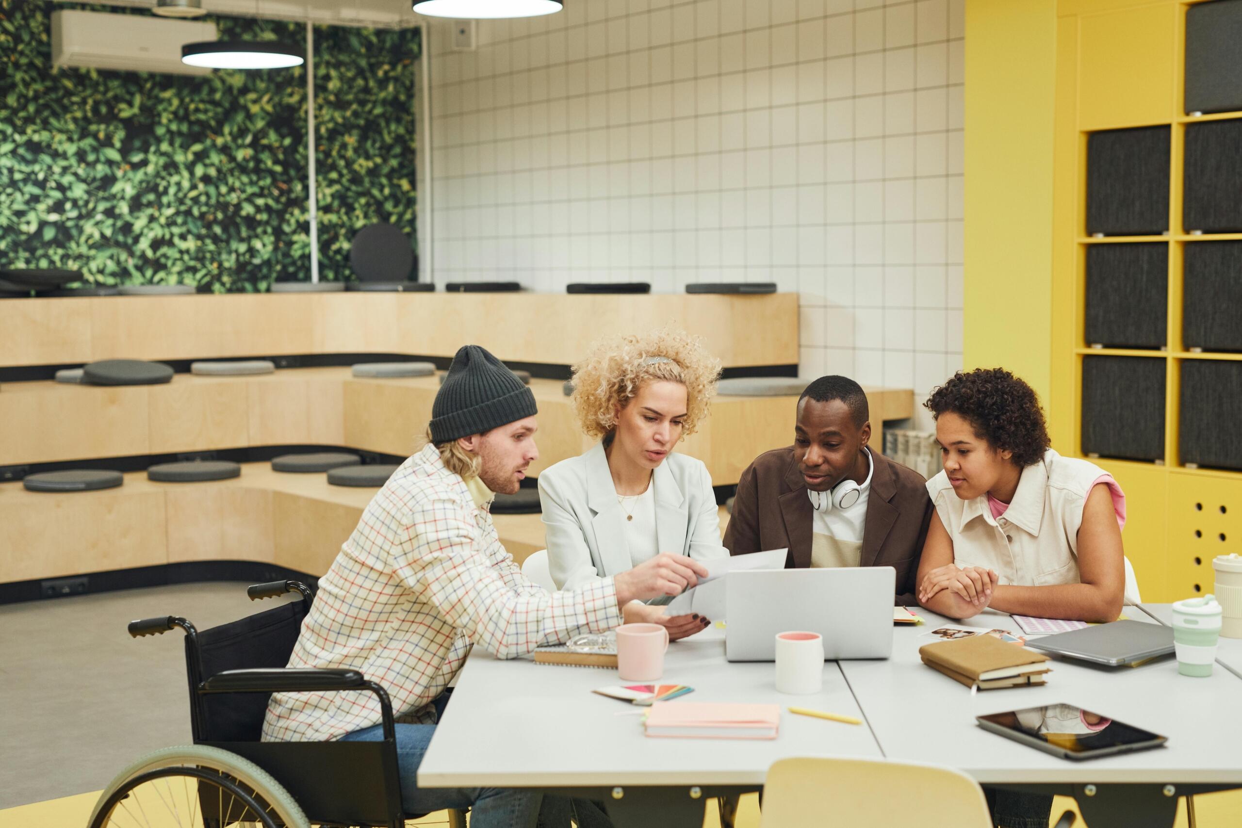 Groep diverse collega’s, waaronder een man in een rolstoel, buigt zich over een laptop aan een tafel in een moderne werkruimte.